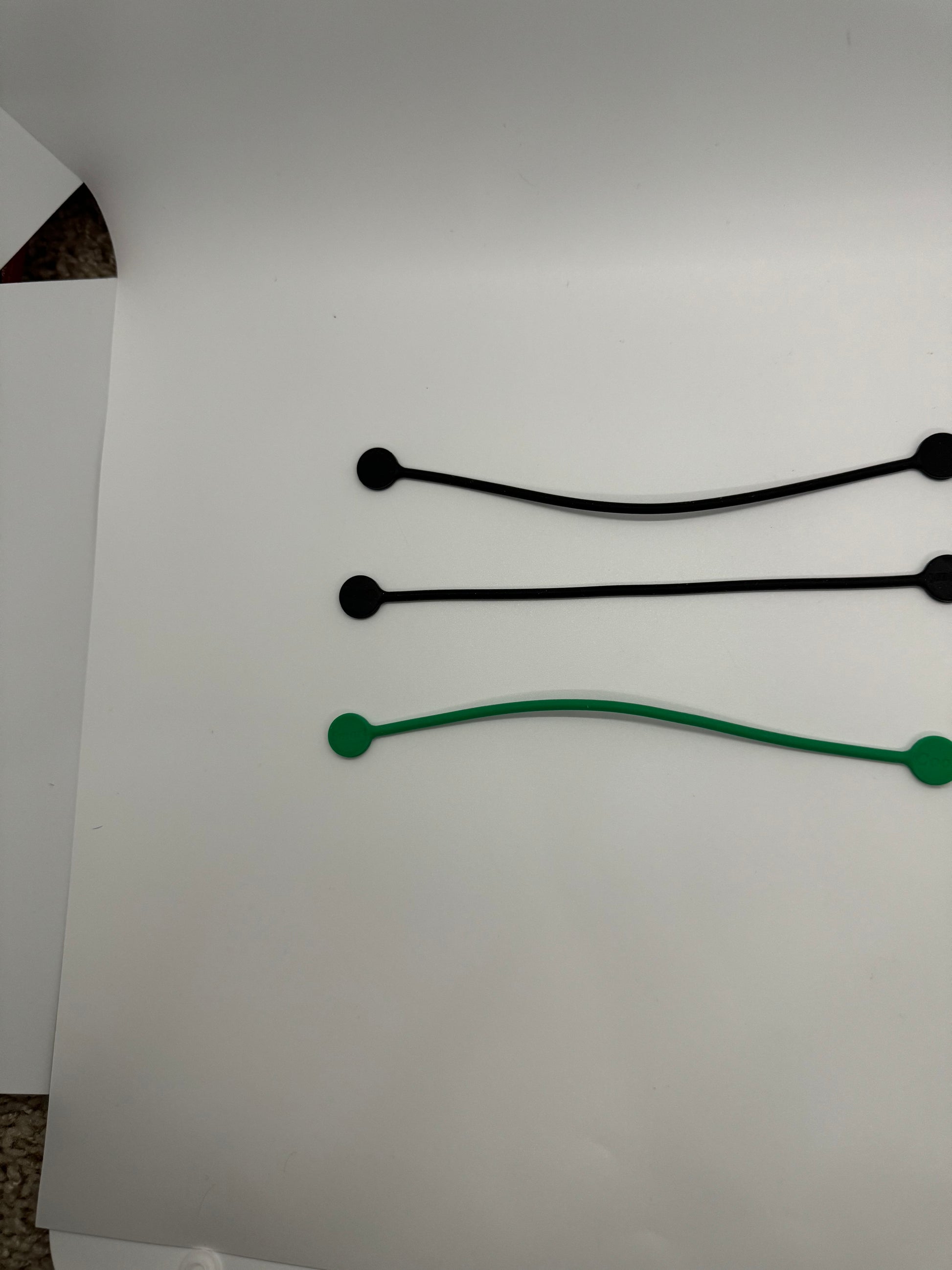 The picture shows three objects on a white background. There are two black bungee cords with balls at each end, placed one above the other. Below them, there is a similar object but in green color. The green one is slightly curved. The background is plain white and there is a small portion of a carpet visible in the bottom left corner.