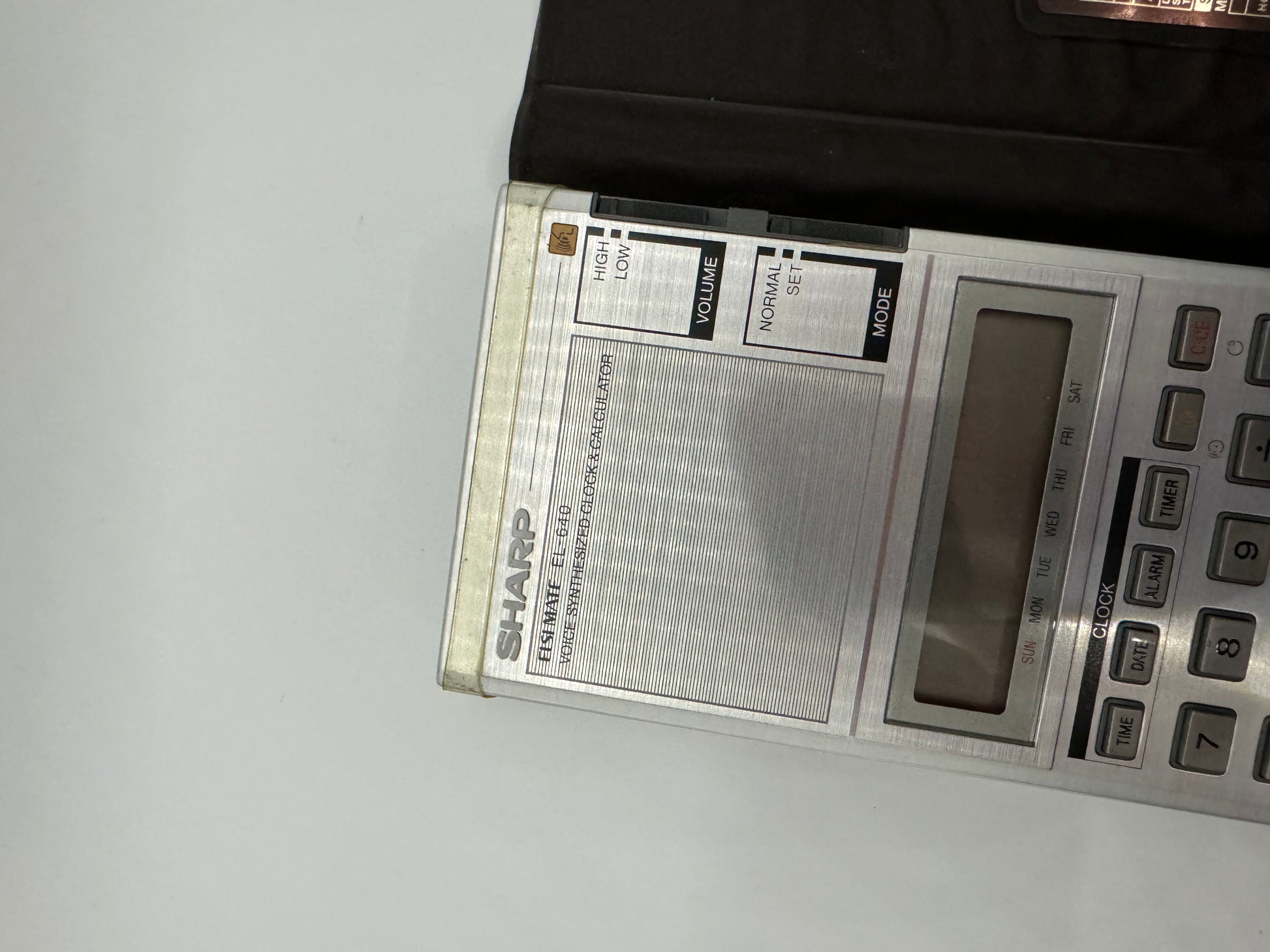 The picture shows a portion of a Sharp electronic device, possibly a calculator or a radio. The device is silver in color. On the left side of the image, there is a black object partially visible. The device has a rectangular screen which is blank. Below the screen, there are several buttons. The buttons are rectangular and grey in color. Some of the buttons have labels such as "CLOCK", "DATA", and numbers from 7 to 9. On the left side of the device, there are two sliders labeled "VOLUME" and "MOD