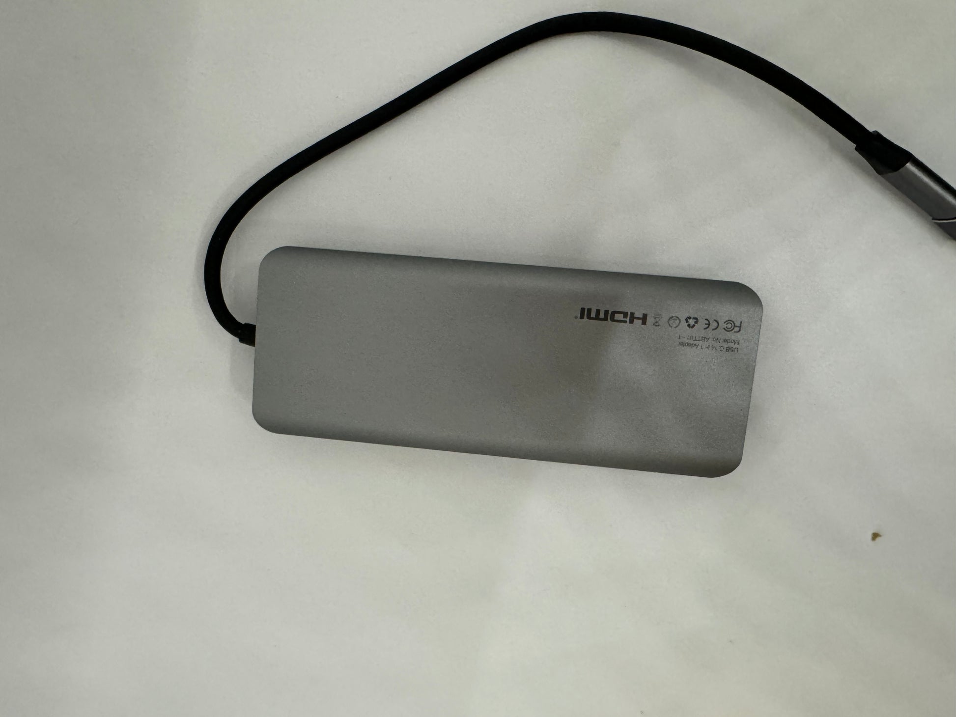 The picture shows a rectangular, grey object with rounded corners. It appears to be a portable electronic device, possibly a power bank or an external hard drive. There is a black cable attached to it. The device has text on it which reads "IWCH" followed by some symbols. Below that, there is some smaller text which is not very clear, but it seems to include "MADE IN CHINA". The background is white with a slight texture.