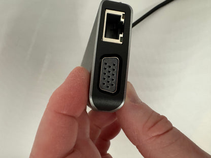 The picture shows a person holding a small electronic device between their thumb and index finger. The device is black and has a rectangular shape with rounded corners. On one side of the device, there is an Ethernet port where you can plug in an Ethernet cable. Next to the Ethernet port, there is a series of small holes arranged in a rectangular pattern, possibly for ventilation or a speaker. The device also has a cable attached to it, but the other end of the cable is not visible in the picture.