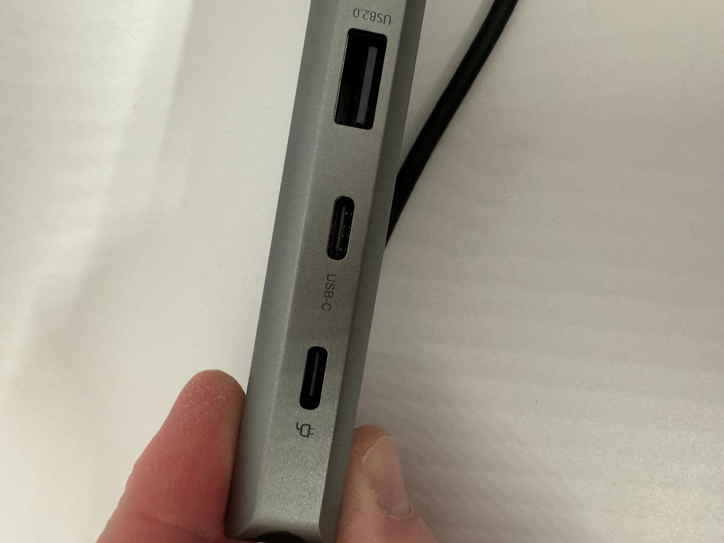 The picture shows a close-up of the side of a silver electronic device, possibly a laptop or a hub. There are two ports visible on the side of the device. The top port is labeled "USB-C" and has a cable plugged into it. The bottom port is smaller and has a symbol next to it that looks like a display monitor. The device is being held by someone's hand, with their thumb visible at the bottom of the picture. The background is a plain white surface.