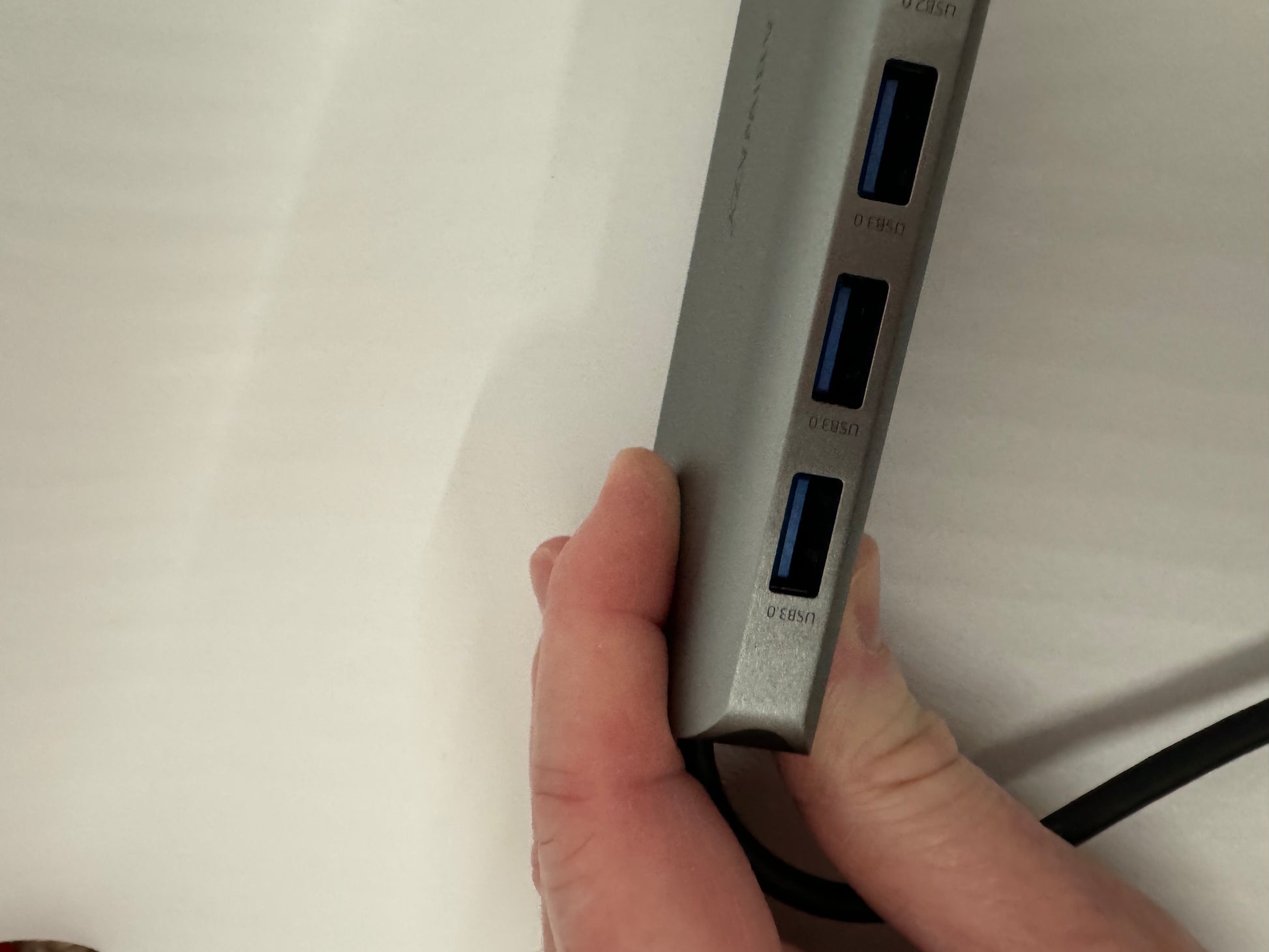 The picture shows a hand holding a gray USB hub. The USB hub has four USB ports, which are vertically aligned. Each port has a label above it that reads "USB 3.0". The background is a plain white surface.