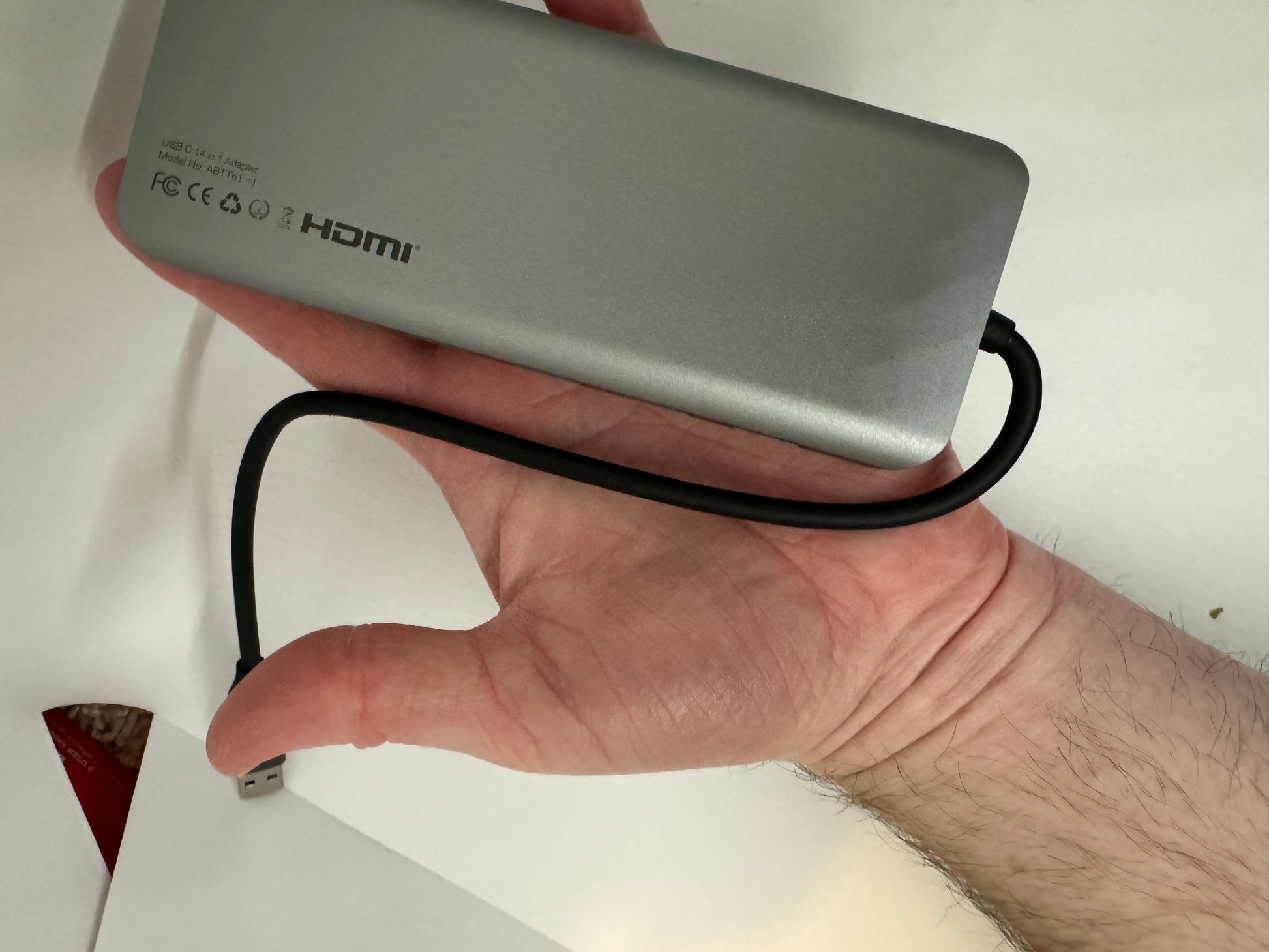 The picture shows a hand holding a rectangular device that appears to be made of metal with a matte finish. The device is silver in color and has a cable attached to it. The cable is black and has a USB connector at the end. On the device, there is text that reads "USB 3.0 to HDMI Adapter" and "Model No: ABT01F". There are also various symbols including "FC", "CE", and the HDMI logo. The background is white and there is a small red object in the bottom left corner. The hand holding the device appe