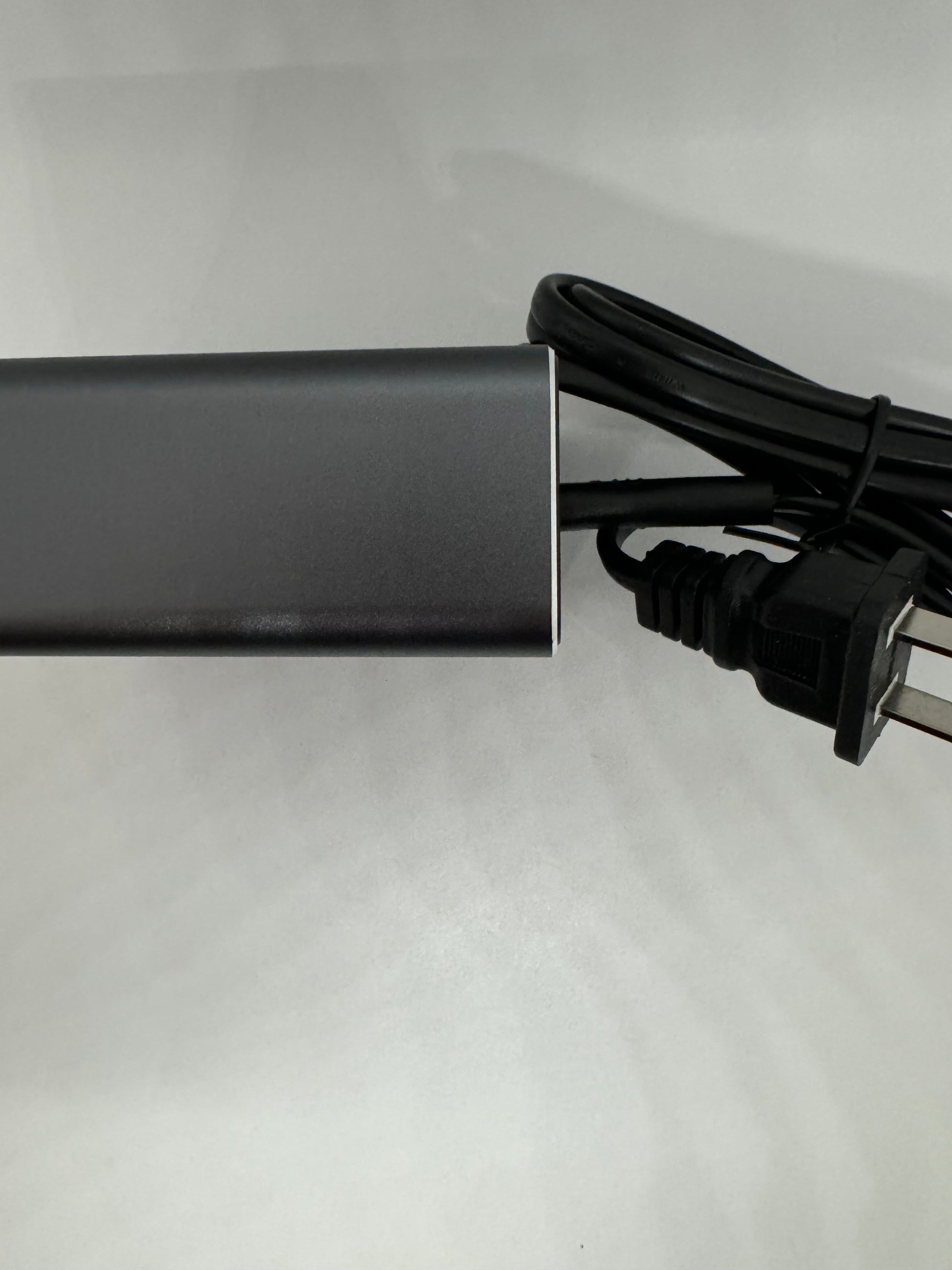 The picture shows a black rectangular object, which appears to be an electronic device, possibly a power bank or an external hard drive. It has a smooth surface and is placed on a white background. There is a black cable with a plug attached to it, coiled up next to the rectangular object. The cable seems to be a power cord or a connector cable. The overall setting is simple and the focus is on the rectangular object and the cable.