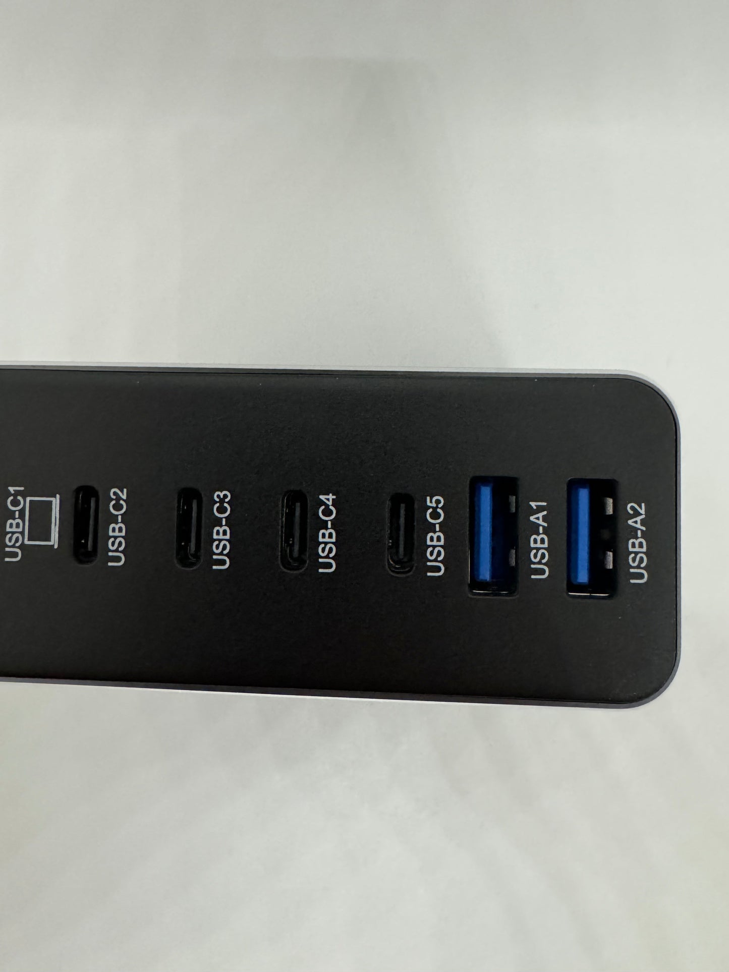 The picture shows a black device with multiple USB ports. The device is rectangular and has a matte finish. There are seven USB ports in total. The ports are labeled from left to right as follows:- USB-C1- USB-C2- USB-C3- USB-C4- USB-C5- USB-A1- USB-A2The first five ports (USB-C1 to USB-C5) are smaller and have a rectangular shape with rounded corners. The last two ports (USB-A1 and USB-A2) are slightly larger and have a rectangular shape. The background is white.