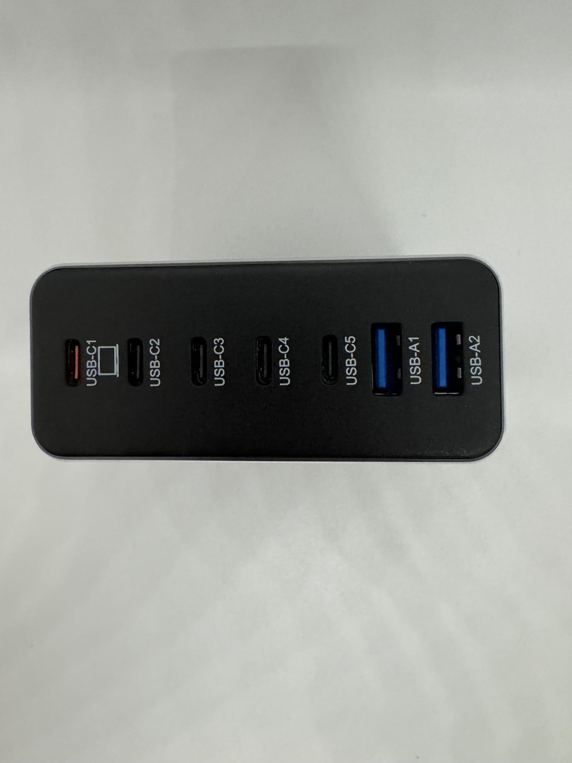 The picture shows a black USB hub with multiple ports. The hub is placed at the bottom of the image against a white background. There are seven ports in total. From left to right, they are labeled as follows:- USB-C 1- USB-C 2- USB-C 3- USB-C 4- USB 3.0 1- USB 3.0 2- USB 3.0 3The USB-C ports are smaller and have a rectangular shape with rounded corners. The USB 3.0 ports are larger and have a rectangular shape. The inside of the USB 3.0 ports is blue.