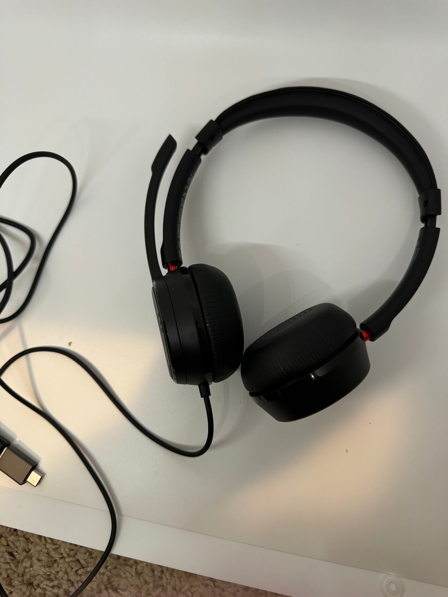 The picture shows a pair of black headphones. The headphones have a cushioned over-ear design and a black adjustable headband. There is a microphone attached to the left side of the headphones. The headphones are connected to a black cable, which has a USB connector at the end. The headphones are placed on a white surface, and there is a beige carpet visible in the bottom part of the picture.