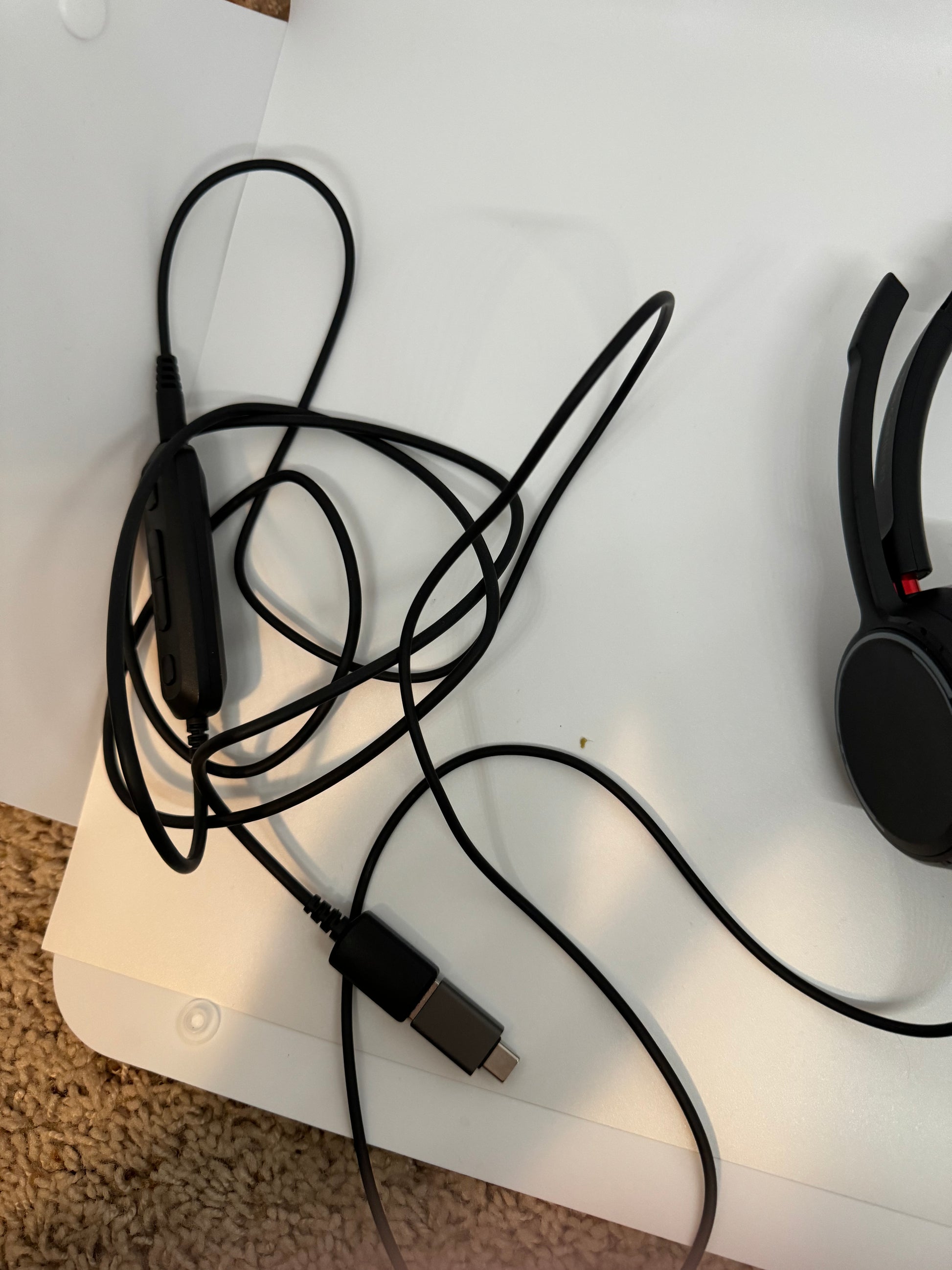 The picture shows a black headset with a long cable. The cable is tangled and has a volume control attached to it. The end of the cable has a USB connector. The headset and cable are placed on a white surface, and the bottom part of the picture shows a beige carpet. The headset appears to have a cushioned earpiece and a red accent on the side.