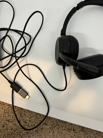 The picture shows a pair of black headphones with a cable attached to them. The headphones have a sleek design with cushioned ear cups. The cable is black and has a USB connector at one end. The headphones are placed on a white surface, and the cable is slightly tangled. There is also a beige carpet visible at the bottom of the picture.