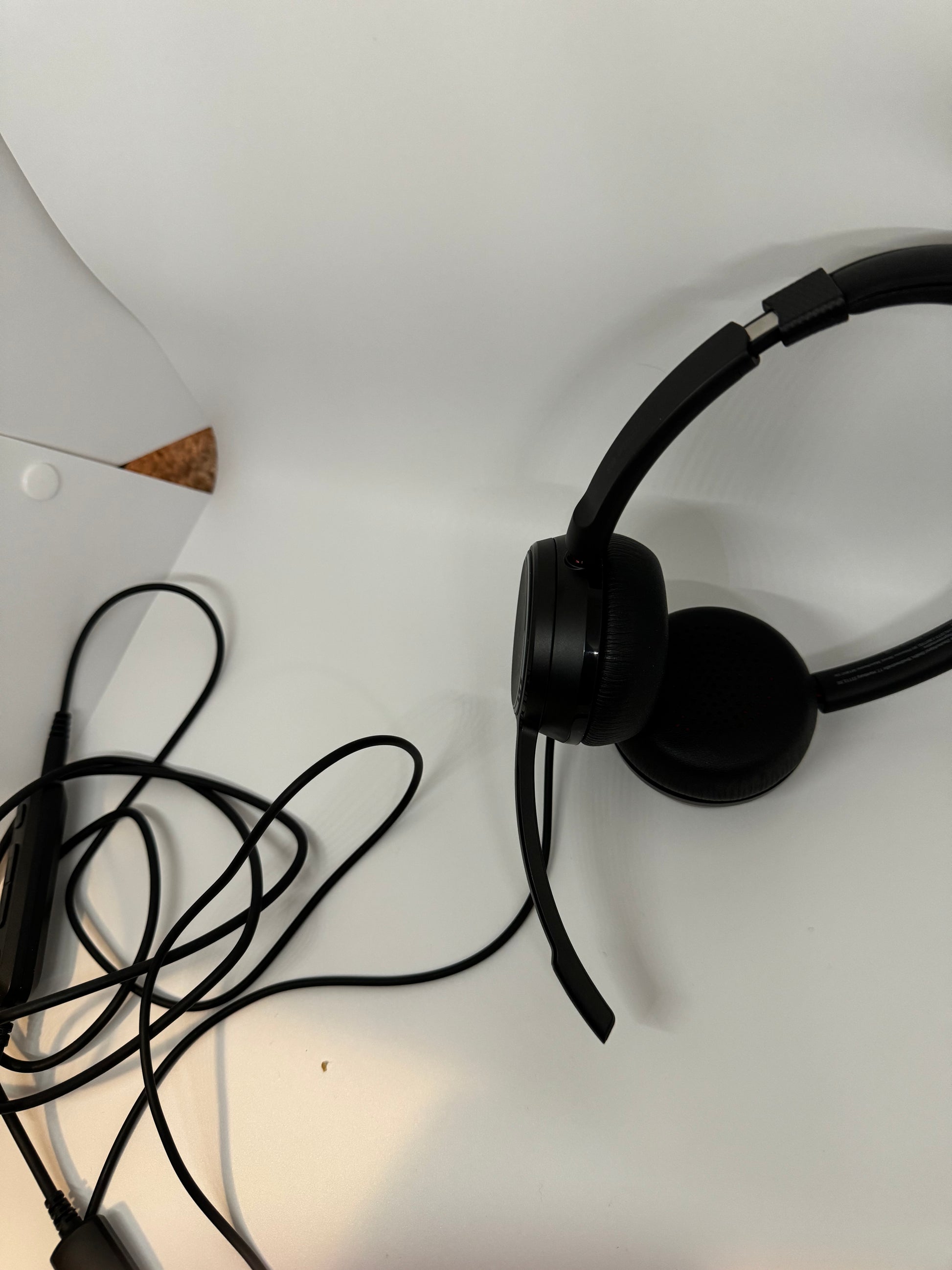 The picture shows a pair of black headphones on a white surface. The headphones have a black cord attached to them. The cord is tangled and has a control module attached to it. The background is mostly white with a small portion of a corkboard visible in the top left corner. There is also a tiny speck or crumb on the white surface near the bottom of the picture.