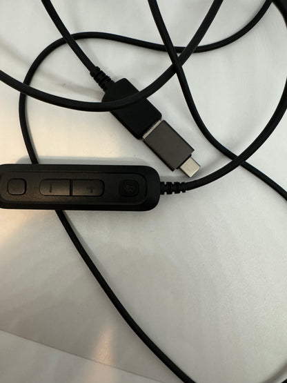 The picture shows a black cable with a USB-C connector on one end. The cable is coiled and laid on a white surface. There is also a small black remote control-like device attached to the cable. The device has two buttons on it, one with a plus sign and the other with a circle. The cable is plugged into this device.