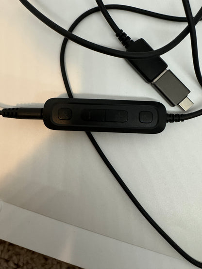 The picture shows a black cable with a control module attached to it. The control module is rectangular and has three buttons on it. From left to right, the first button has a symbol that looks like a microphone, the middle button is longer and rectangular, and the third button has a symbol that looks like a headset. The cable also has a USB-C connector at the end. The background is a white surface.