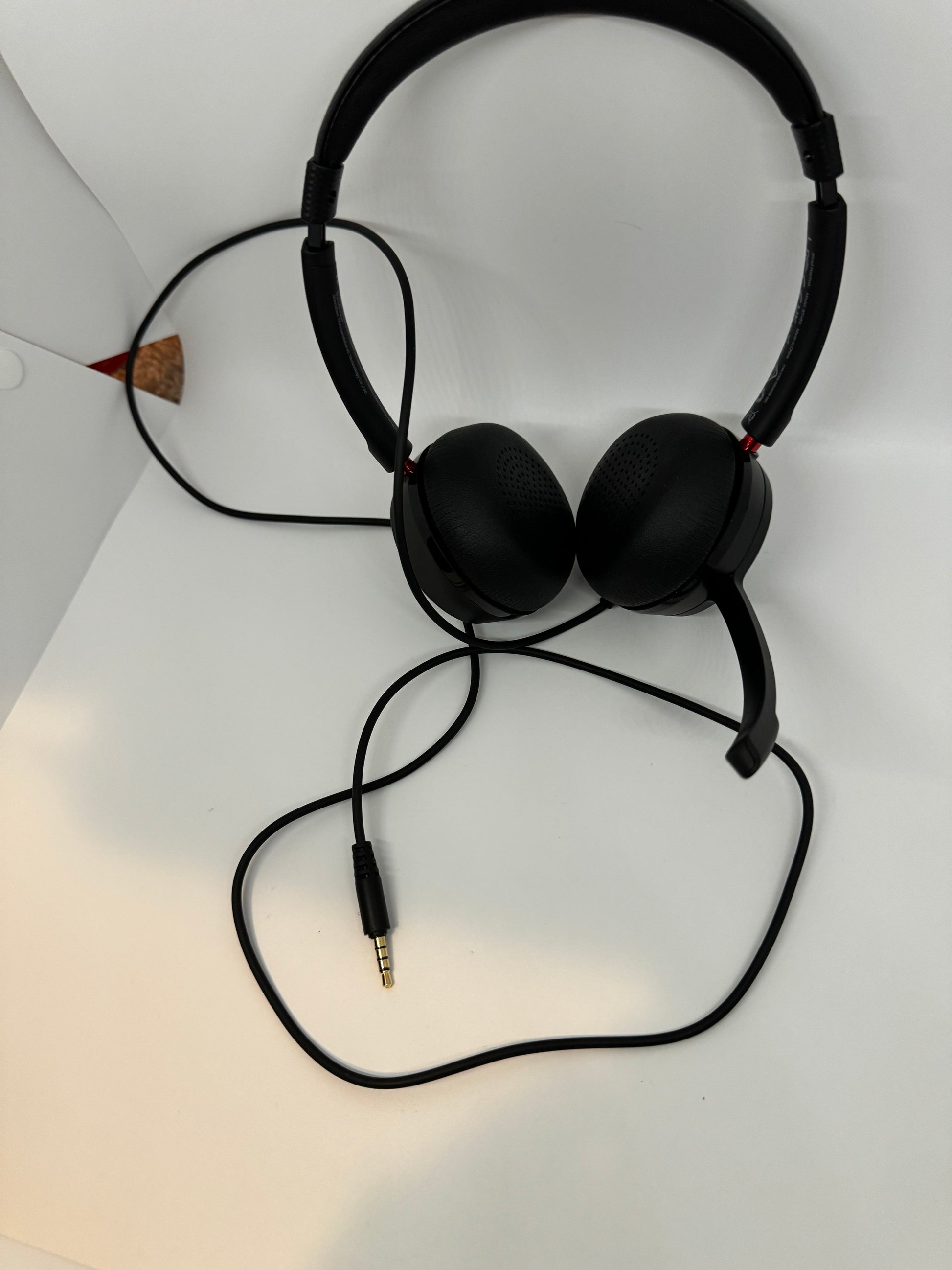 The picture shows a pair of black headphones. The headphones have a black headband and two black ear cups with perforated patterns. The ear cups are connected to the headband with a red accent. There is a black cable attached to the headphones, which has a 3.5mm audio jack at the end. The background is white, and there is a small part of something that looks like a cork material with a red tip in the top left corner, but it's mostly out of frame.