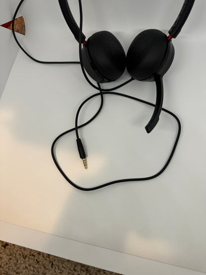 The picture shows a pair of headphones. The headphones are black with a red accent near the ear cups. They have a cushioned headband and perforated ear cups. The headphones are placed on a white surface, and the cable is visible, leading to a 3.5mm audio jack. The background also shows a beige carpet.