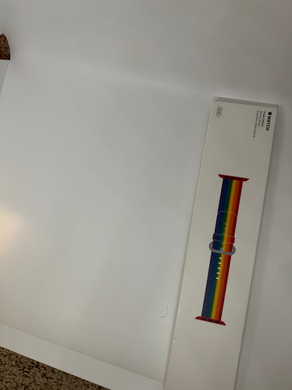 The picture shows a white rectangular box lying on a white surface. The box is positioned diagonally from the top right corner to the bottom left corner of the image. On the box, there is an image of a watch band with a rainbow color scheme. The colors on the band are arranged in horizontal stripes, with red at the top, followed by orange, yellow, green, blue, and purple. Above the image of the watch band, there is text that reads "WATCH" in black capital letters. Below that, there is smaller text