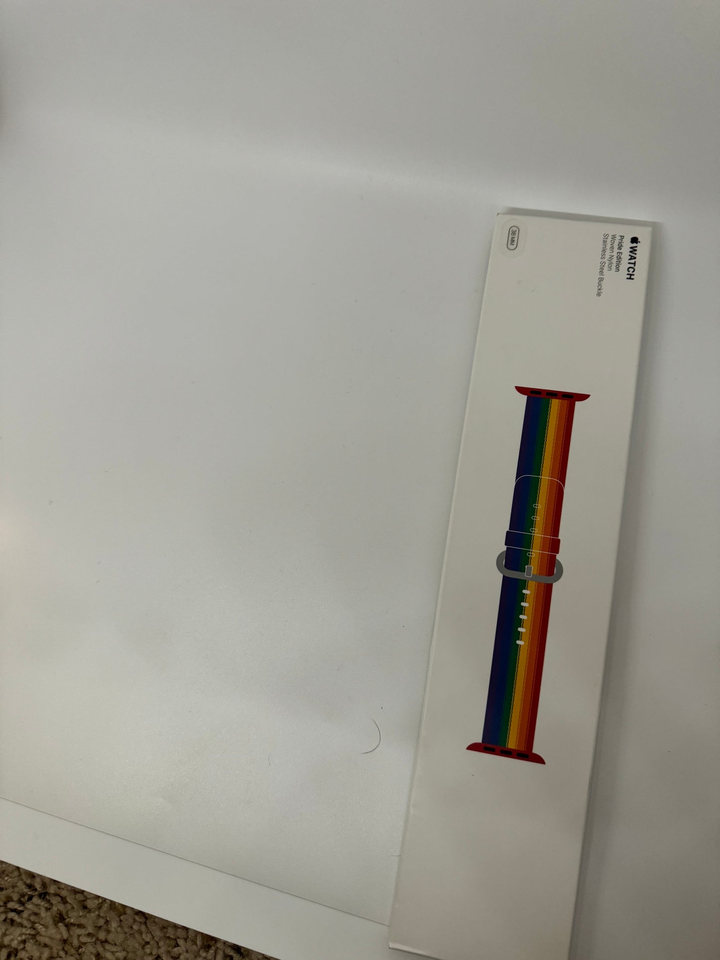 The picture shows a white rectangular box placed on a white surface. The box is positioned vertically and is partially visible on the right side of the image. On the box, there is an image of a watch band that has a rainbow color scheme. The colors on the band are arranged in vertical stripes and include red, orange, yellow, green, blue, and purple. Above the image of the watch band, there is some text which is not fully visible, but the word "WATCH" can be seen. The background is plain white and 