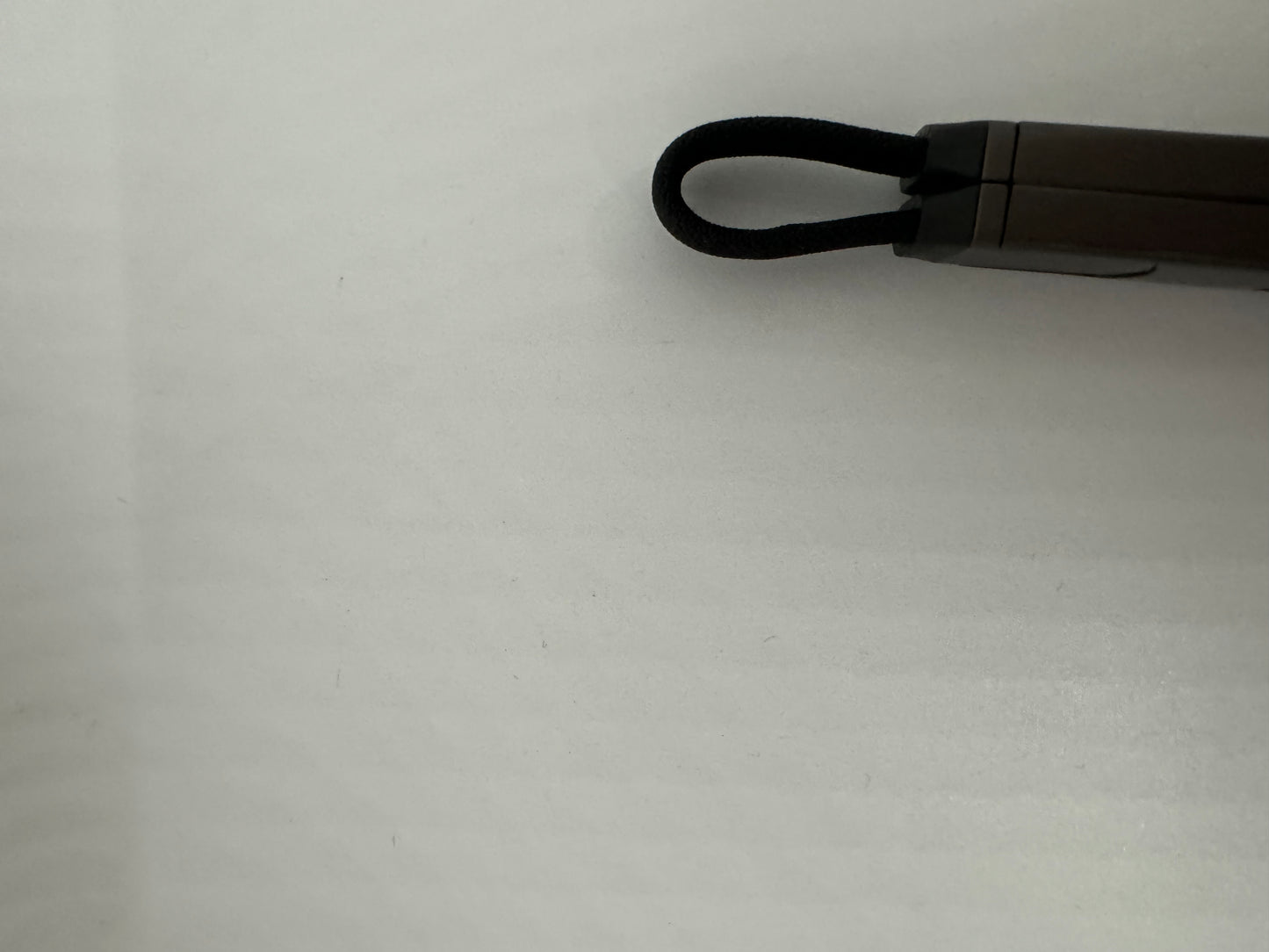 The picture shows a close-up of a white surface with a small object in the top right corner. The object appears to be a pen or a similar tool. It has a dark gray or black body with a looped fabric strap attached to its end. The strap is black and looks like it's made of a woven material. The pen or tool is lying horizontally, and the background is plain white with some shadows and texture.