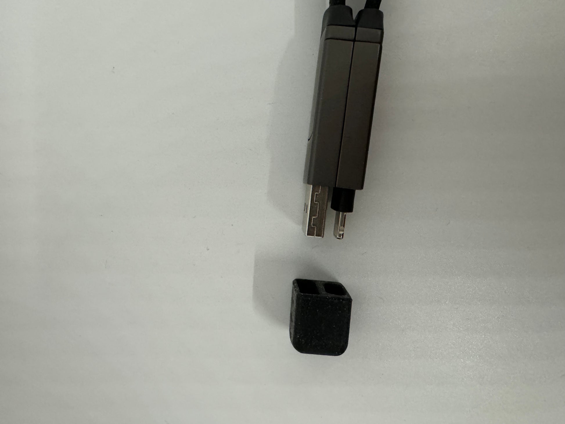 The picture shows two objects on a white surface. On the right side, there are two black cables with connectors at the end. The connectors are rectangular and metallic with a small protrusion on one side. On the bottom right corner, there is a small black object that looks like a cap or cover. It is rectangular with one of the shorter sides open. The surface of this object appears to be textured.