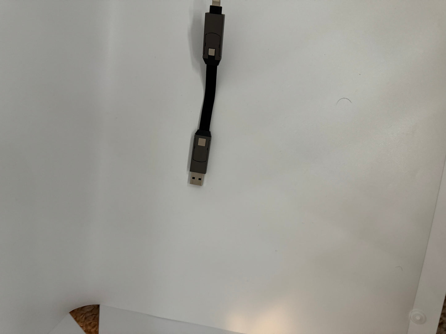 The picture shows a USB cable against a white background. The cable is black and flat. On the left side of the image, the cable has a standard USB connector, and on the right side, it has a smaller connector, possibly a micro USB or USB-C. The cable is entering the frame from the top left corner and exiting through the bottom right corner. There is also a small, curved black line or object on the white background near the center of the image.