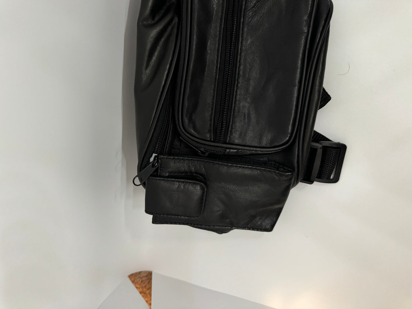 The picture shows a black leather bag. The bag appears to be a small sling bag or waist pouch. It has multiple zippers and pockets. There is a zipper at the top and a smaller zipper in the front. The bag also has a black strap with a plastic buckle, which is likely used to secure it around the waist or across the body. The bag is placed on a white surface.