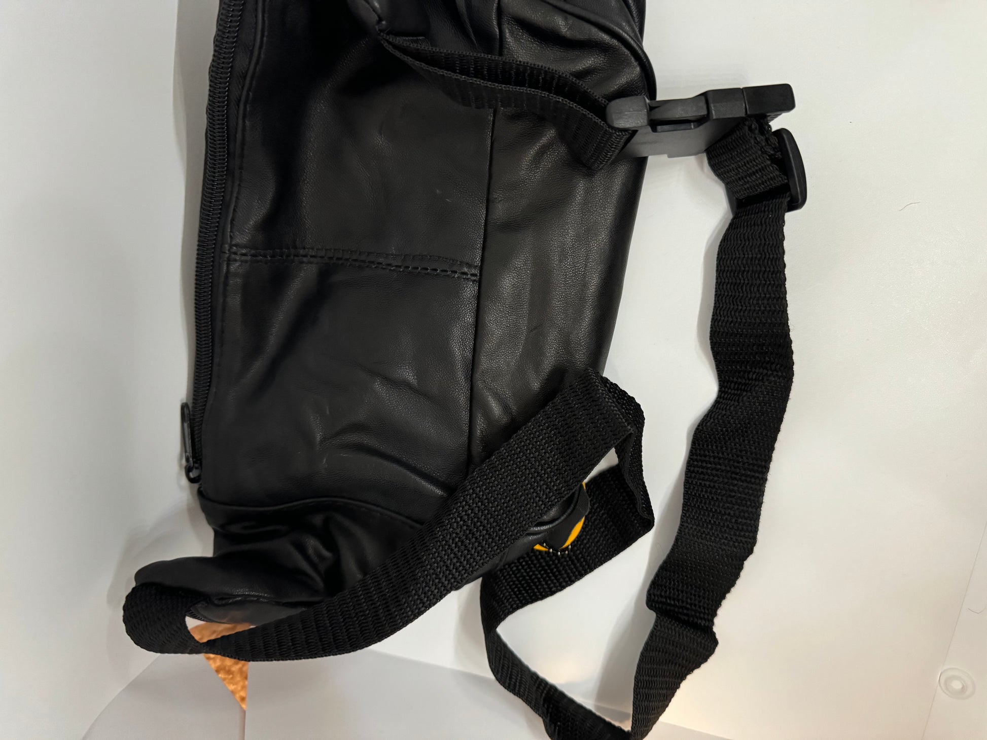 The picture shows a black bag, possibly made of leather or a similar material. The bag has a zipper on the side. There is also a black strap attached to the bag with a plastic buckle. The strap is made of a woven material and looks sturdy. The bag is placed on a white surface.