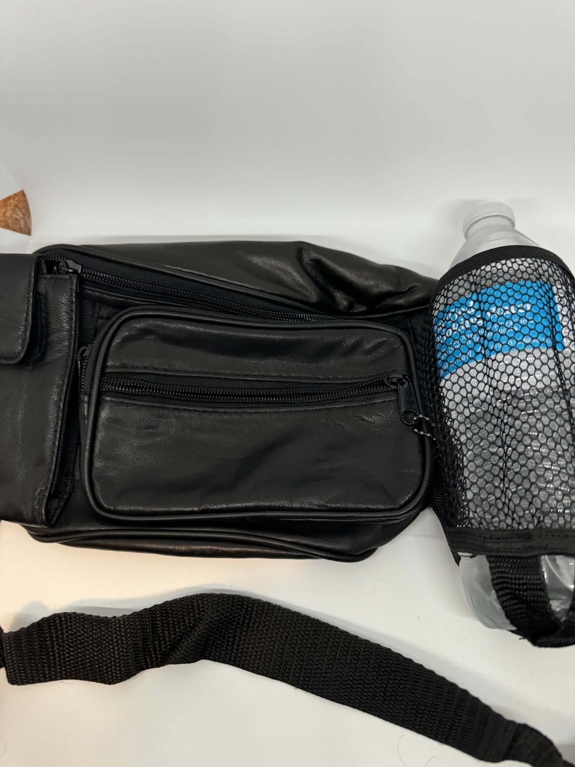 The picture shows a black waist bag (fanny pack) with a water bottle next to it. The waist bag is made of black leather and has multiple zippered compartments. The strap of the waist bag is also visible and it is made of a black woven fabric. The water bottle is in a black mesh holder and has a blue honeycomb pattern on it. The background is plain white.