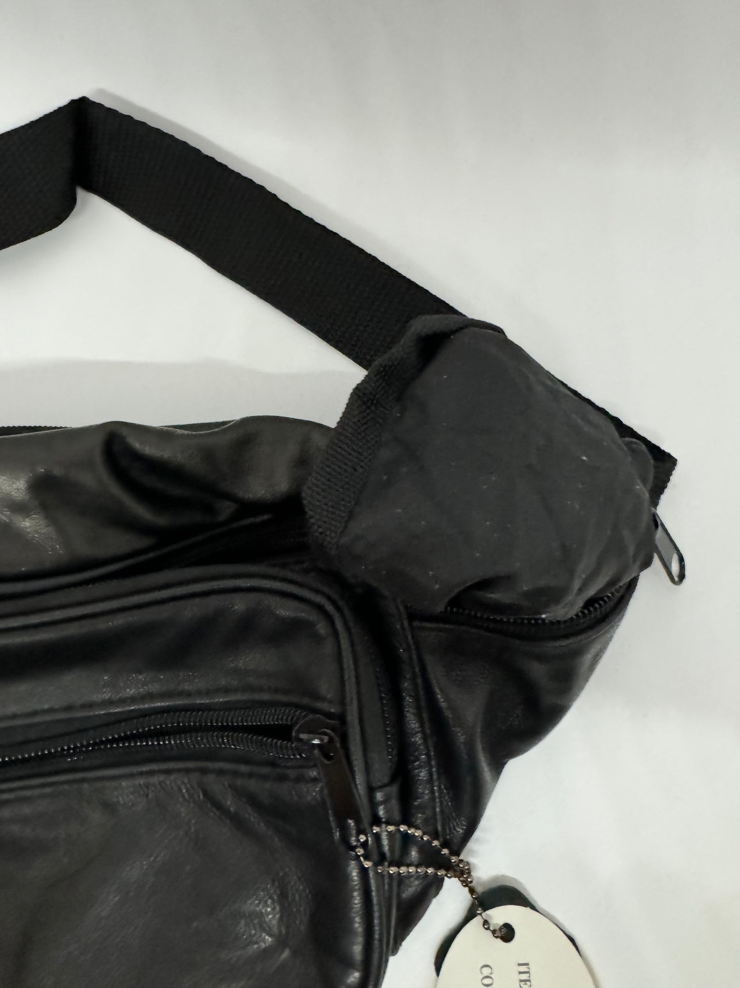 The picture shows a black bag, possibly made of leather or a similar material. The bag has a zipper on the top and a smaller zipper on the side. There is a black strap attached to the bag, which is made of a woven material. The strap is thick and seems sturdy. There is also a tag attached to the bag with a ball chain, but the text on the tag is not visible in the picture. The background is plain white.