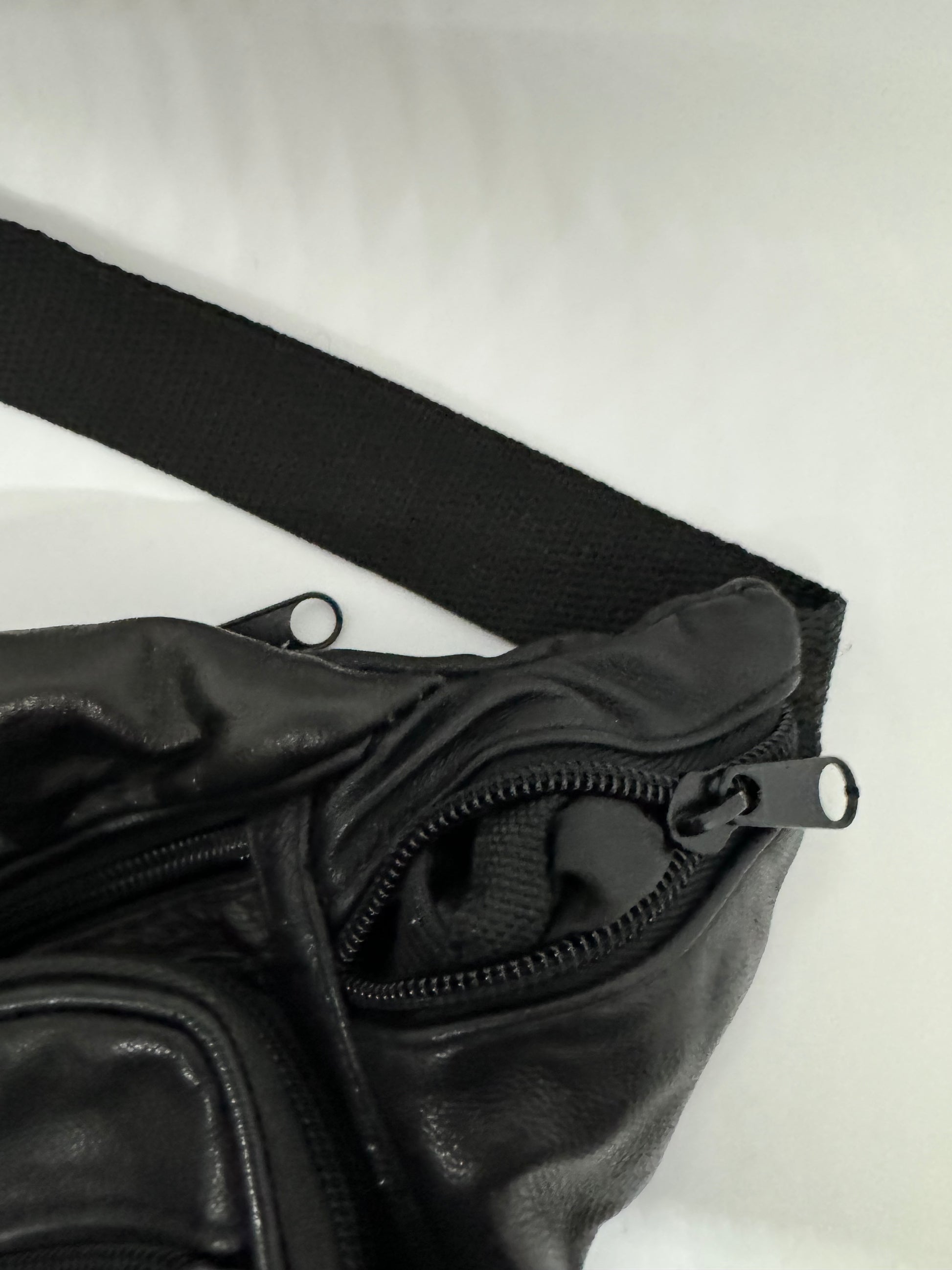 The picture shows a black item made of a material that appears to be leather or a similar fabric. It has a zipper with a metal pull tab. The zipper is partially open, revealing a black inner lining. There is also a black woven strap in the picture, which is lying next to the leather-like item. The background is white.
