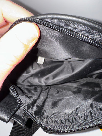 The picture shows a close-up of someone's hand holding the edge of a black fabric item, possibly a bag or pouch. The person's thumb is visible, and they are holding the edge of the item to show the zipper and the fabric. The zipper is black with fine teeth. The fabric appears to be black and has a slight sheen to it. There is also a small white tag visible inside the item. The background is not very clear, but it seems to be a plain surface.