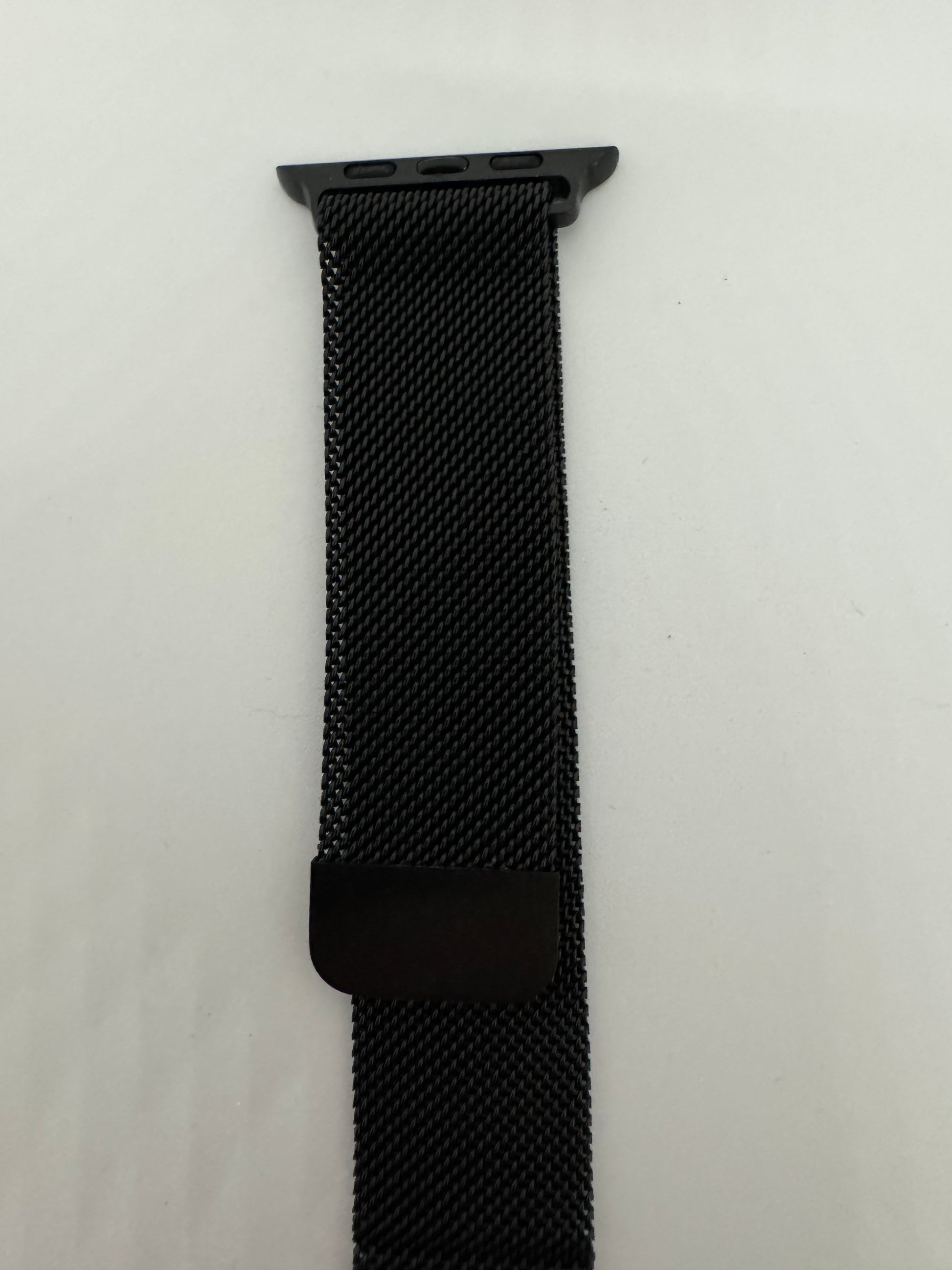 The picture shows a black watch band. It appears to be made of a tightly woven mesh material. The top of the band has a dark grey or black attachment with three small holes, which is likely used to connect it to a watch. The band is lying on a white surface. There is also a small black rectangular piece attached to the band, possibly for adjustment or decoration.