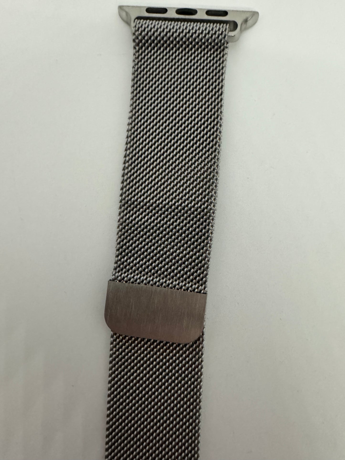 The picture shows a watch band. It is made of a tightly woven metal mesh, giving it a flexible and smooth texture. The color of the band is silver. At the top of the band, there is a silver attachment with three small holes on each side, which is used to connect the band to a watch. About halfway down the band, there is a rectangular metal clasp that is used to adjust the size and secure the band around the wrist. The background of the picture is white.