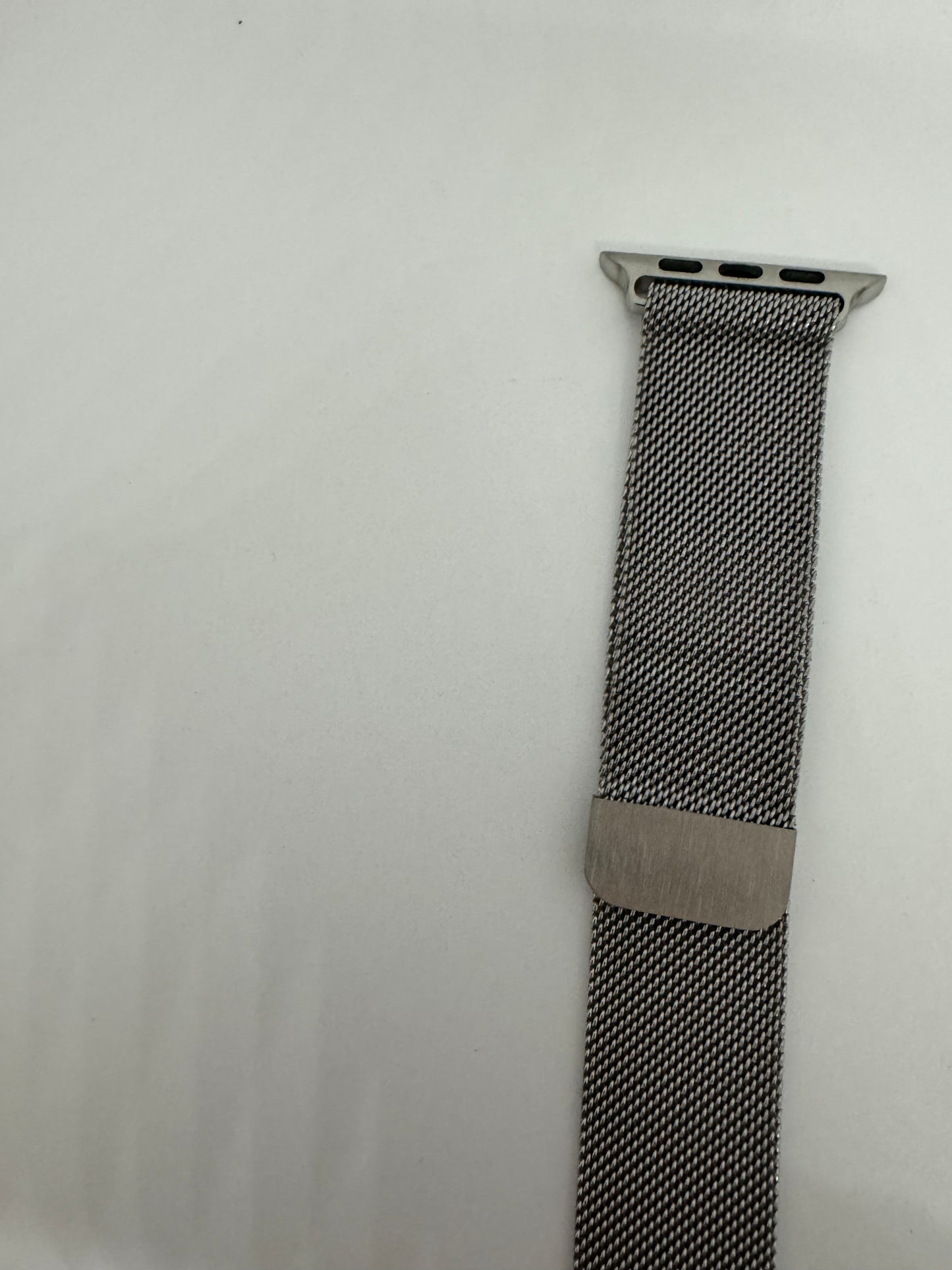 The picture shows a watch band against a white background. The band is made of a tightly woven metal mesh, giving it a sleek and modern appearance. The color of the band is a dark metallic, possibly stainless steel. The top part of the band has a connector with three small holes, which is likely used to attach it to a watch. There is also a metal clasp on the band, which is used to secure it around the wrist. The band appears to be new or in very good condition.