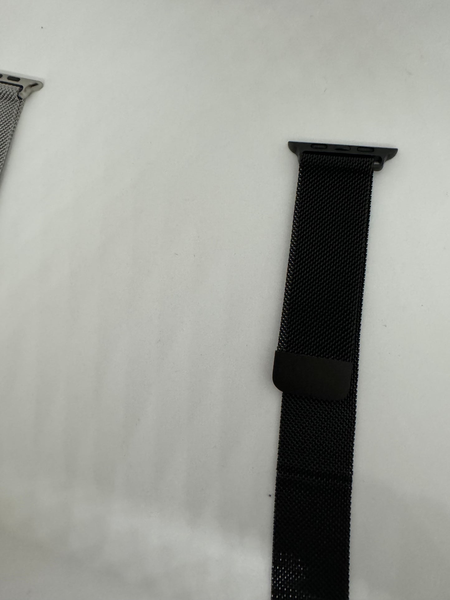 The picture shows two watch bands on a white background. On the left side, there is a small portion of a silver-colored watch band visible. On the right side, there is a black watch band with a dark grey attachment piece at the top. The black band appears to be made of a woven material and has a small black loop near the top. The background is plain white.