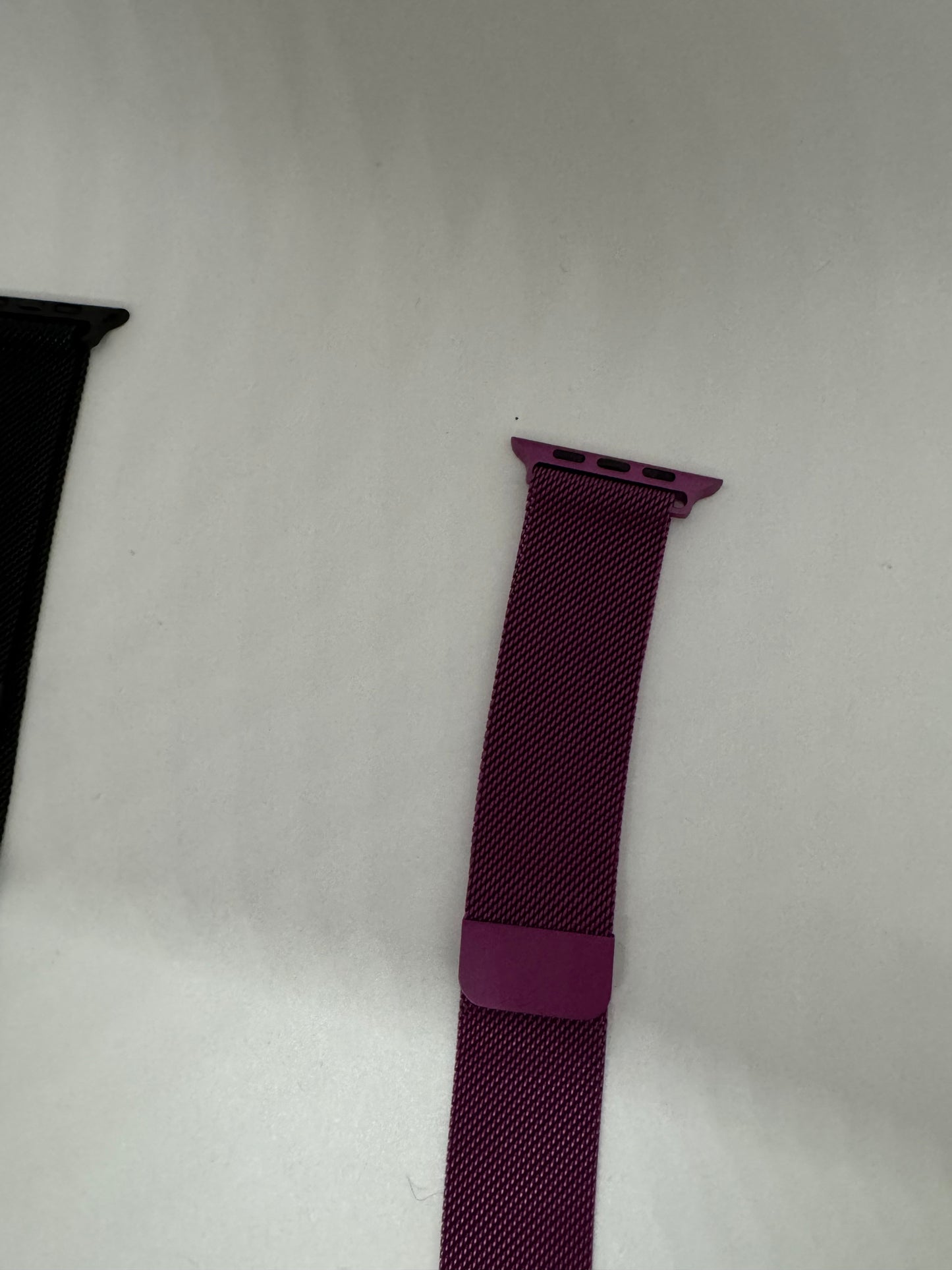 The picture shows two watch bands lying on a white surface. The one on the left is partially visible and appears to be black. The one on the right is more visible and is a vibrant purple color. The purple watch band has a textured pattern and a metal attachment with three holes at the top, which is likely used to connect it to a watch.