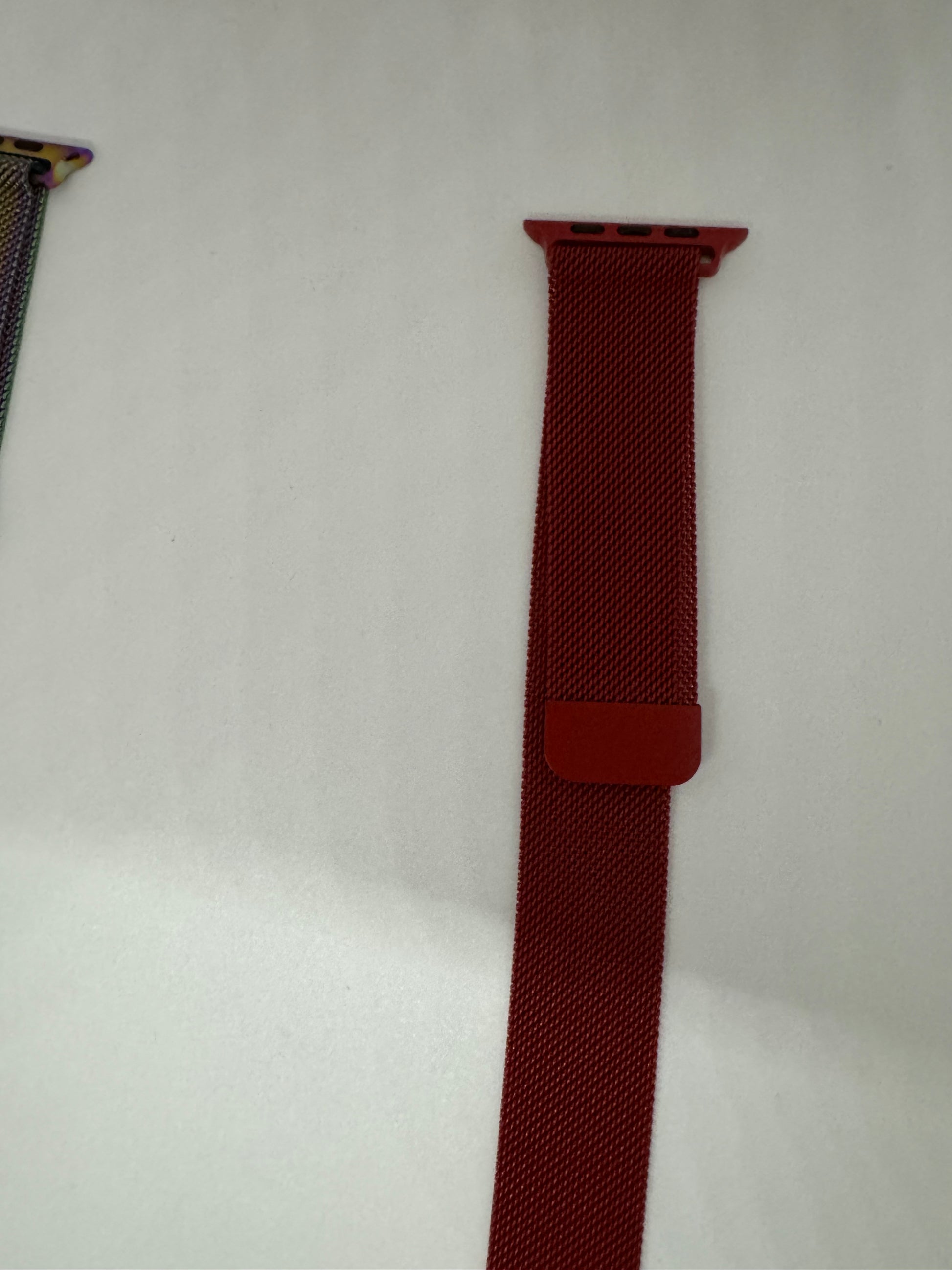 Be My AI: The picture shows two watch bands on a white surface. On the left side, there is a small portion of a watch band visible with a multicolored pattern. On the right side, there is a red watch band. The red watch band has a woven texture and is attached to a connector that is used to attach it to a watch. The connector is also red and has two small black pins. There is also a small red loop on the band.