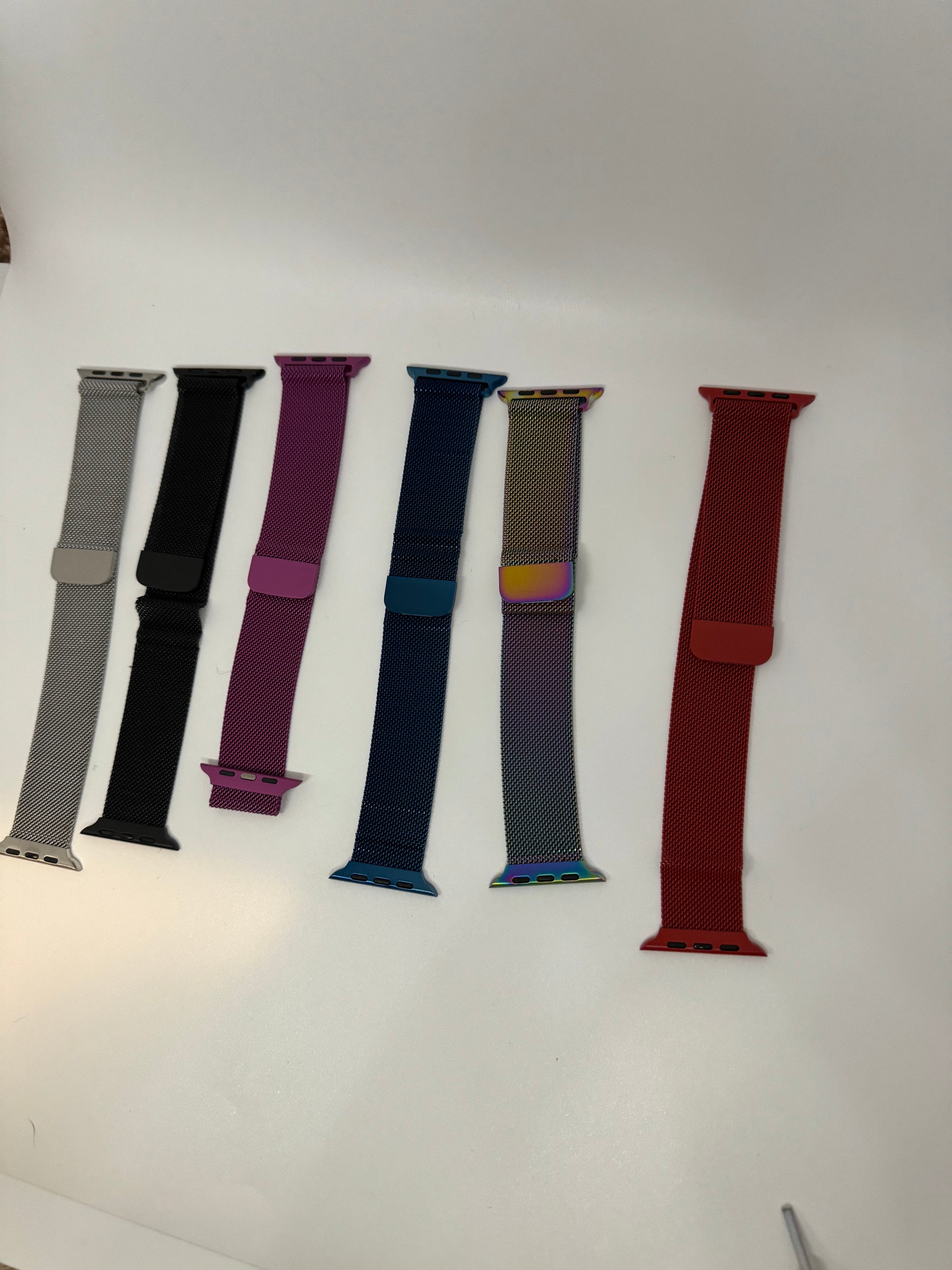 The picture shows six watch bands laid out horizontally on a white surface. From left to right:1. The first band is a silver mesh.2. The second band is solid black.3. The third band is a vibrant magenta color.4. The fourth band is a deep blue.5. The fifth band has a gradient pattern, starting with a golden brown at the top, transitioning to a pinkish-purple in the middle, and ending with a teal color at the bottom.6. The last band on the right is a solid dark red color.Each band has metal