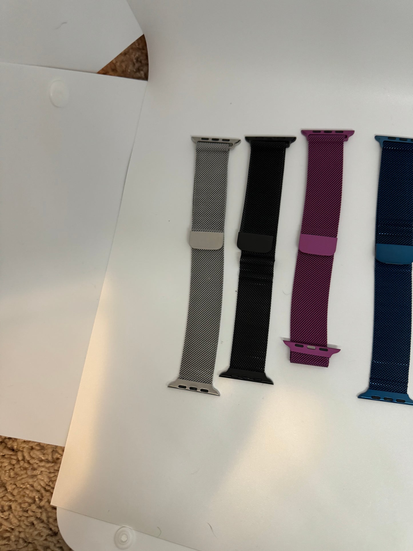 The picture shows four watch bands laid out on a white surface. From left to right, the first band is silver with a metallic mesh texture. The second band is black and appears to have a smooth texture. The third band is a vibrant pink color with a similar texture to the black band. The fourth band is blue with a slightly darker shade at the bottom. The bands have attachments at the ends, likely for connecting to a watch face. The background has a white surface with a small portion of a beige carpe