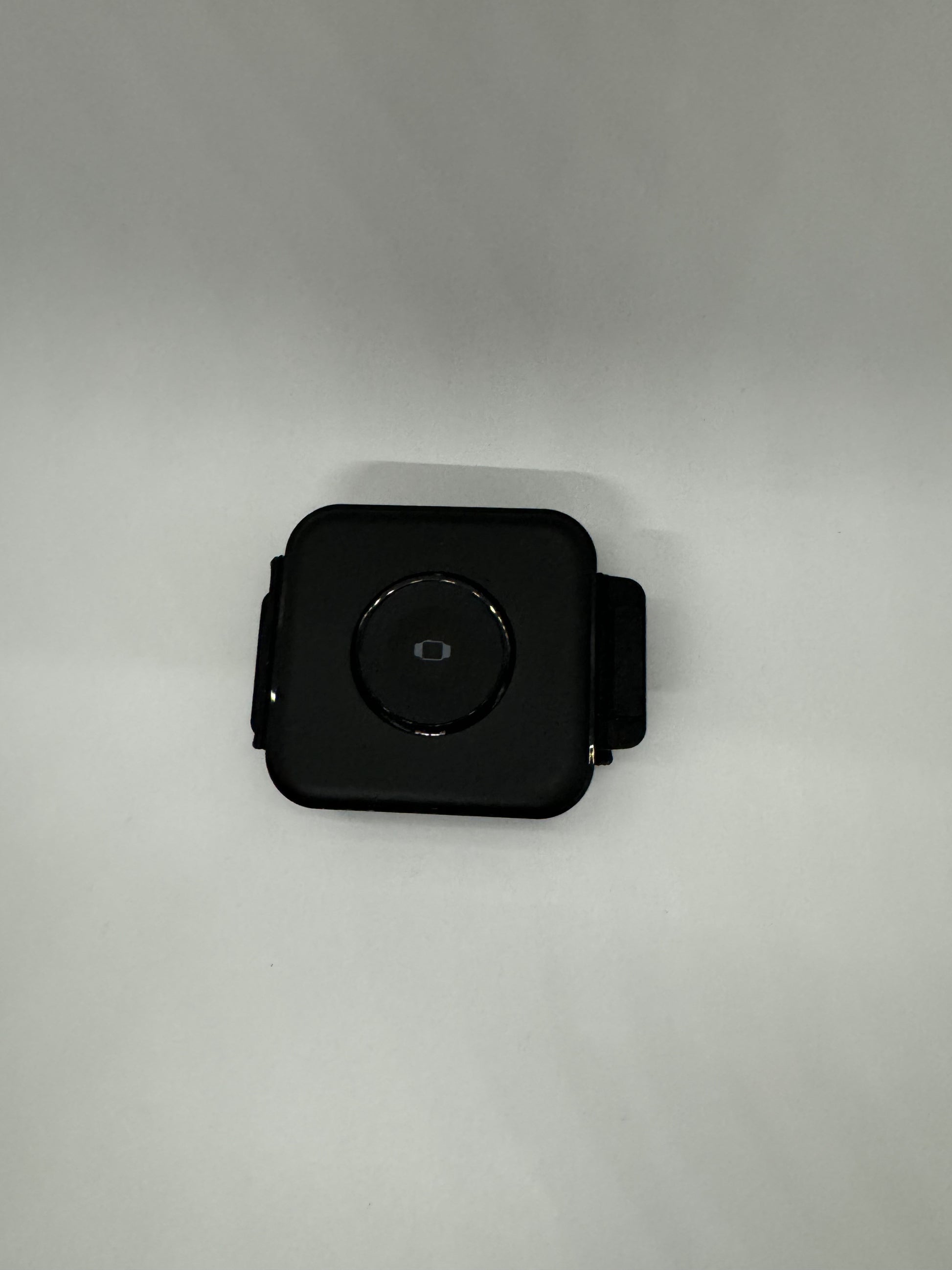 The picture shows a small black square object with rounded corners. It appears to be some sort of electronic device. There is a circular button or dial in the center of the object. The background is plain white. The object is positioned at the bottom of the image.