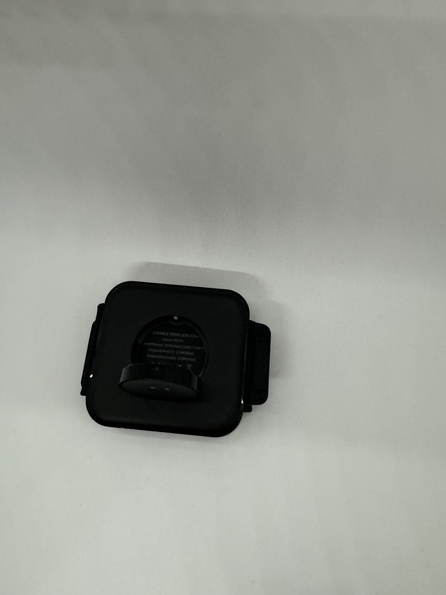 The picture shows a small black object on a white background. The object appears to be a square-shaped device with rounded corners. It has a clip on the back and some text written on it. The text reads: "SQUARE WIRELESS CHARGER, Model: MC-02A, Input: 5V-2A, Output: 5W". There is also a circular indentation in the center of the device, which seems to be the area where you would place a phone or other device for wireless ch