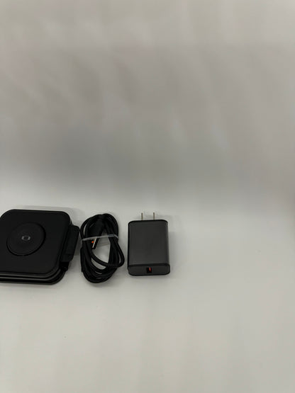 The picture shows three items on a white surface with a light grey background. 1. On the left, there is a black square-shaped object with rounded corners. It appears to be a stack of small square devices, possibly hard drives. The top one has a circular indentation in the center.2. In the middle, there is a black cable coiled up. It seems to be a standard USB cable.3. On the right, there is a black rectangular object with a plug at the top, likely a charger or adapter. It has a red mark on o