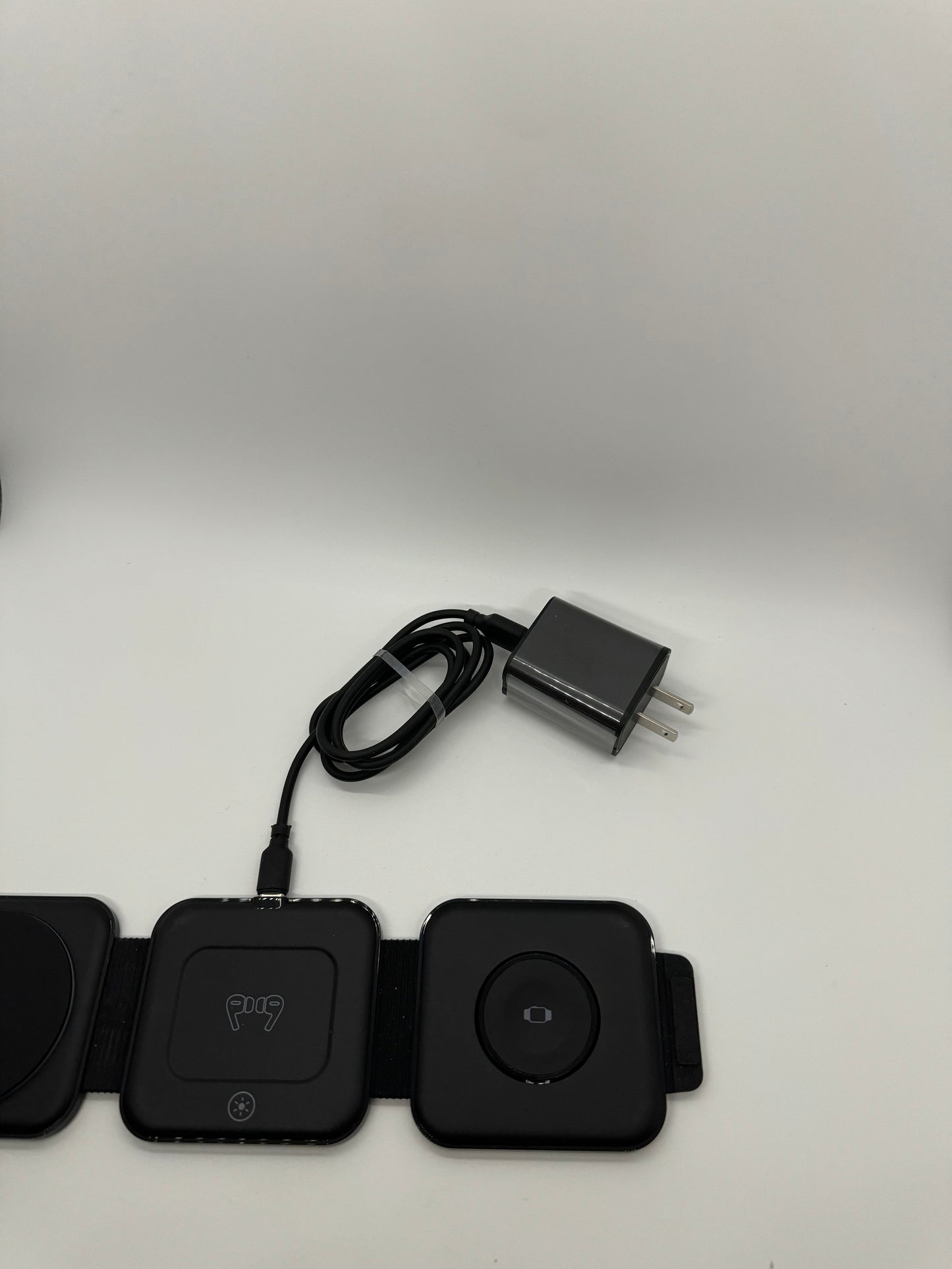 The picture shows three black square devices lying next to each other on a white surface. The device in the middle has a cable attached to it. Above the devices, there is a black power adapter with a two-prong plug and a cable wrapped around it. The device on the left has a circular indentation in the center. The middle device has a logo that looks like a dog's face and a small circular button below the logo. The device on the right has a circular shape in the center with a small square inside i