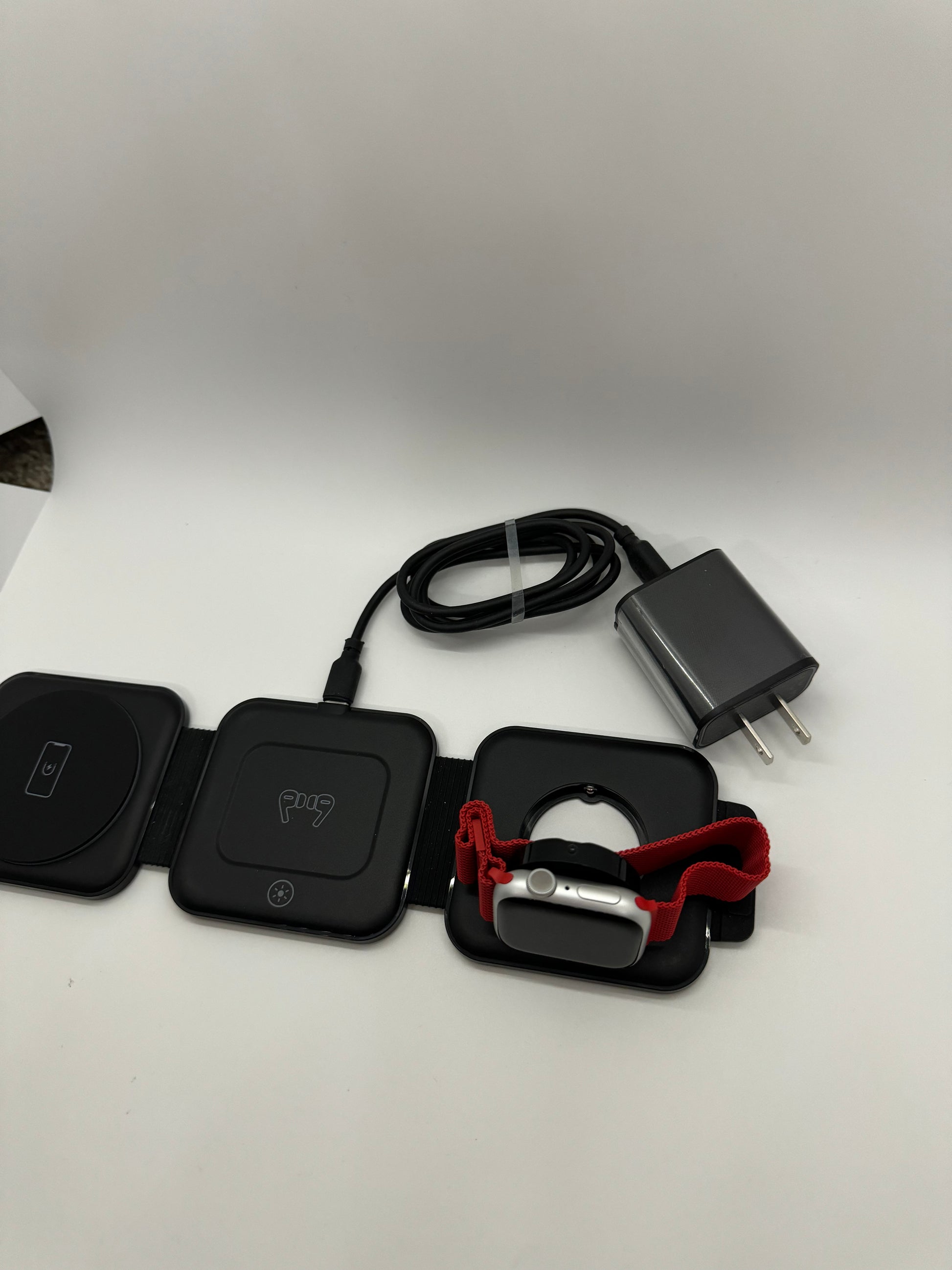 Be My AI: The picture shows several items on a white surface. From left to right:

1. Two black square-shaped devices with rounded corners. They appear to be some sort of electronic devices. The one on the left has a power symbol on it, and the one on the right has the letters "PQI" and a Bluetooth symbol.

2. A black cable coiled up.

3. A gray power adapter with two prongs for a US electrical outlet.

4. A small black device with a red fabric strap. The device looks like it could be a fitness tracker or a