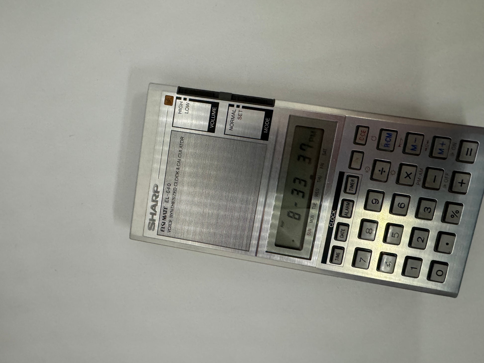 The picture shows a Sharp brand calculator. The calculator is silver and has a rectangular shape. On the left side, there is a speaker grill and above it, there is a switch labeled "Volume" with options "High" and "Low". The brand name "Sharp" is written below the speaker grill. On the right side of the calculator, there is a display screen showing some numbers and letters. Below the screen, there is a set of buttons including numbers 0-9, mathematical operation buttons such as plus, minus, multip