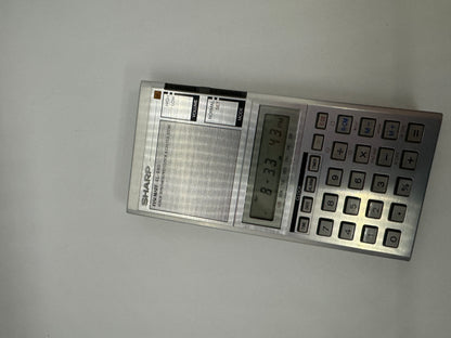 The picture shows a Sharp brand calculator. The calculator is silver and rectangular in shape. On the left side, there is a speaker with a volume control slider above it. The speaker has a grid pattern and the volume control has labels "low" at the bottom and "high" at the top. On the right side of the calculator, there is a digital display screen showing the numbers "13131313". Below the screen, there are buttons for different functions and calculations. The buttons are square and grey in color