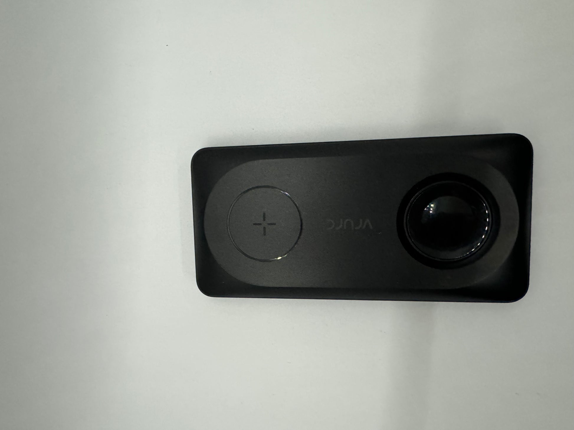 The picture shows a rectangular device that is black in color. On the left side of the device, there is a circular button with a plus sign in the middle. On the right side of the device, there is a round lens or sensor. Below the button, the word "DJI" is written in white letters. The device is placed on a white surface.