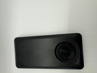 The picture shows a rectangular object with rounded corners. It is black in color and appears to be made of plastic. On the left side of the object, there is a circular indentation with a plus sign in the middle. On the right side, there is a circular opening or lens. The background is plain and white.