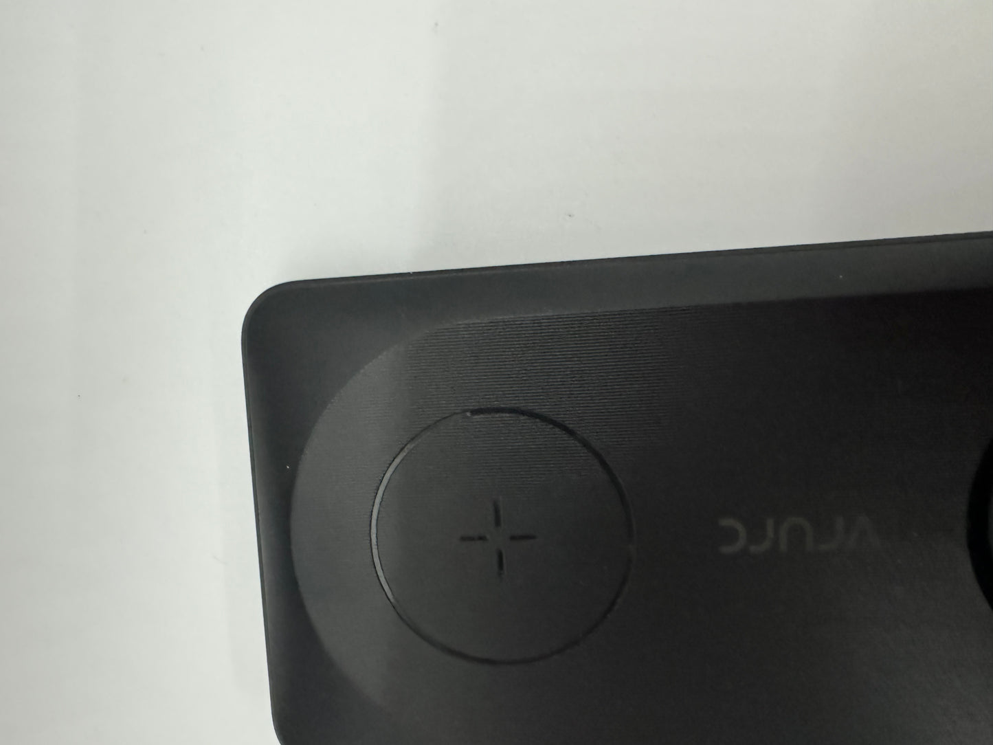 The picture shows a close-up of a dark gray or black object with a matte finish. On the object, there is a circular button with a plus sign on it. The button appears to be for volume control. The word "Anker" is embossed in the same color as the object, making it slightly difficult to see. The background is plain white. The object seems to be some kind of electronic device, possibly a speaker.