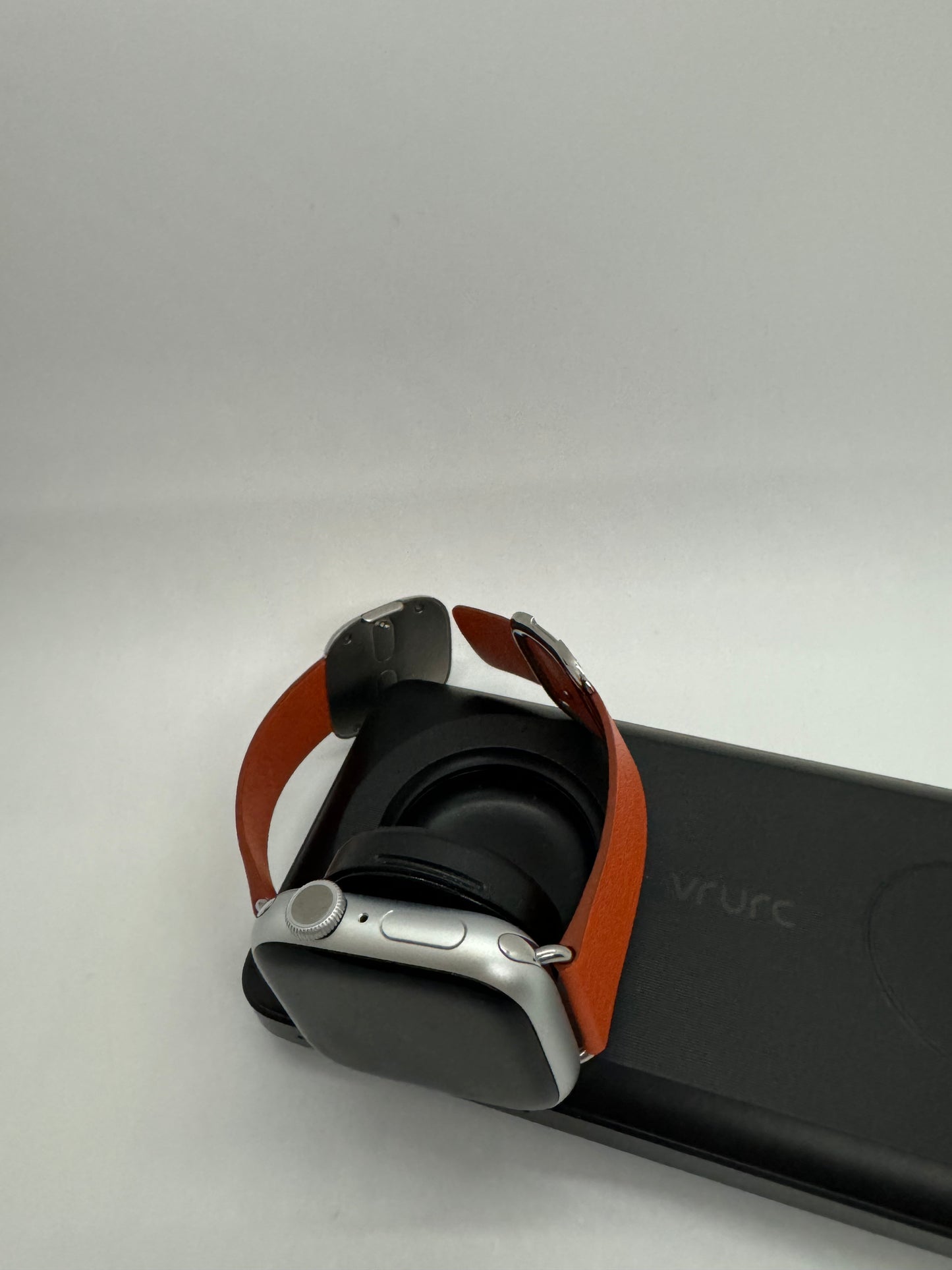 The picture shows a smartwatch with a brown leather strap. The watch is placed on top of a black rectangular object that appears to be a case or a box. The smartwatch has a silver body with a round dial and a button on the side. The black case or box has the word "vrurc" written on it in lowercase letters. The background of the picture is plain white.