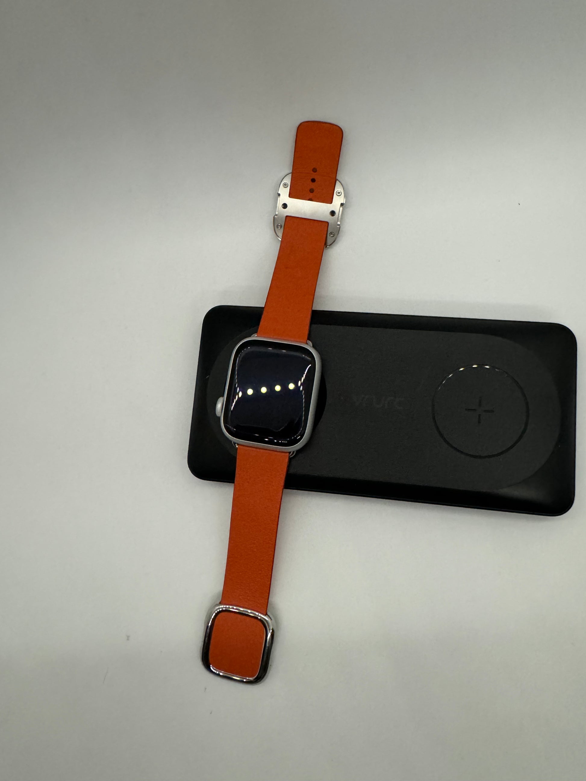 The picture shows a smartwatch with an orange leather strap. The watch face is square with rounded corners and appears to be turned off or in sleep mode as the screen is black. The strap has a silver buckle and there are two loops to secure the strap. The watch is placed on top of a black rectangular object that seems to be a wireless charger. The charger has a plus sign on the top right corner. The background of the picture is plain white.