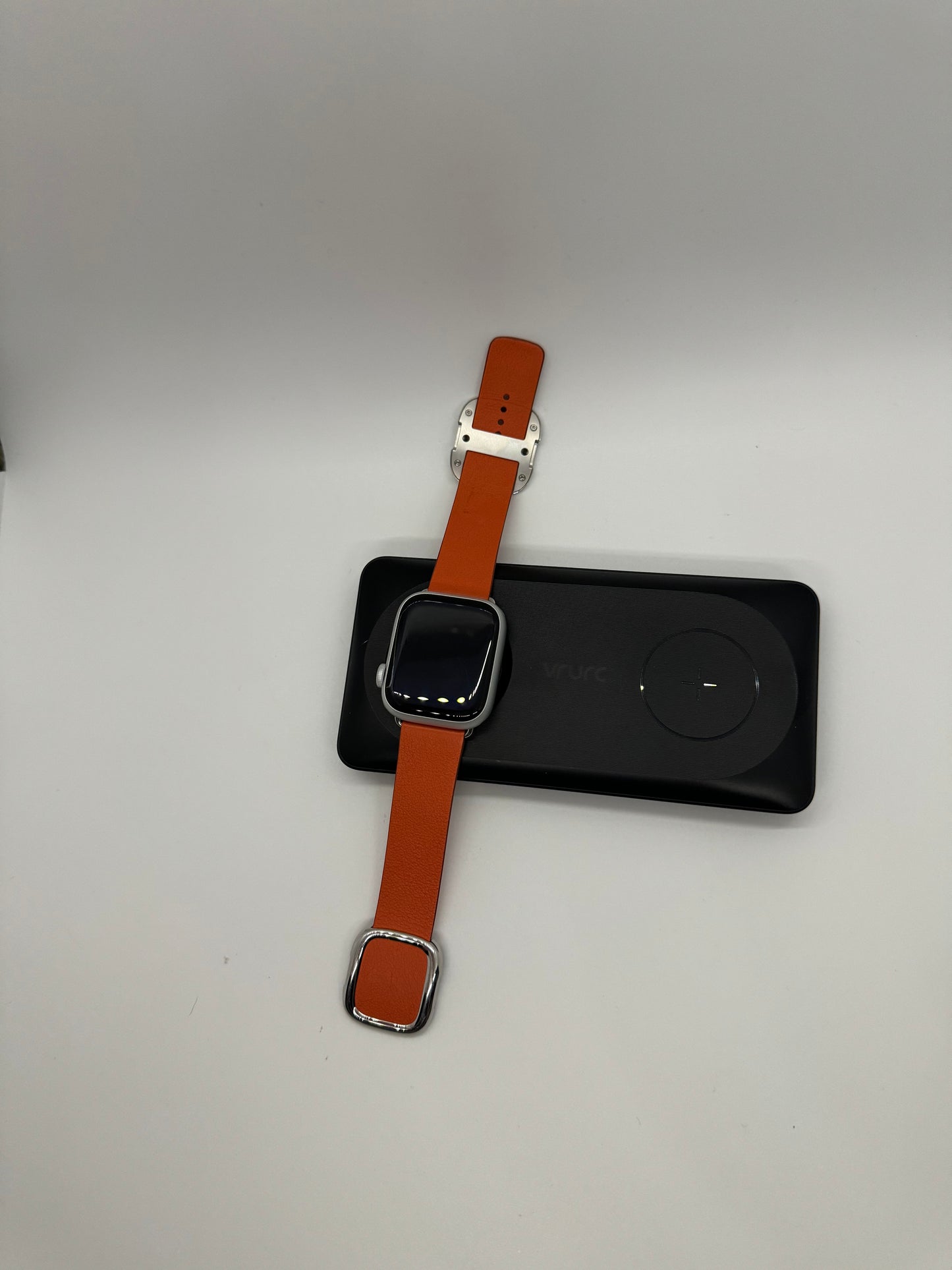 The picture shows a smartwatch with an orange leather strap. The smartwatch is placed on a black rectangular charging pad. The charging pad has a circular indentation where the watch is placed for charging. The watch has a square face with rounded corners and a black screen. The strap has a silver buckle and loops. The background is plain white.